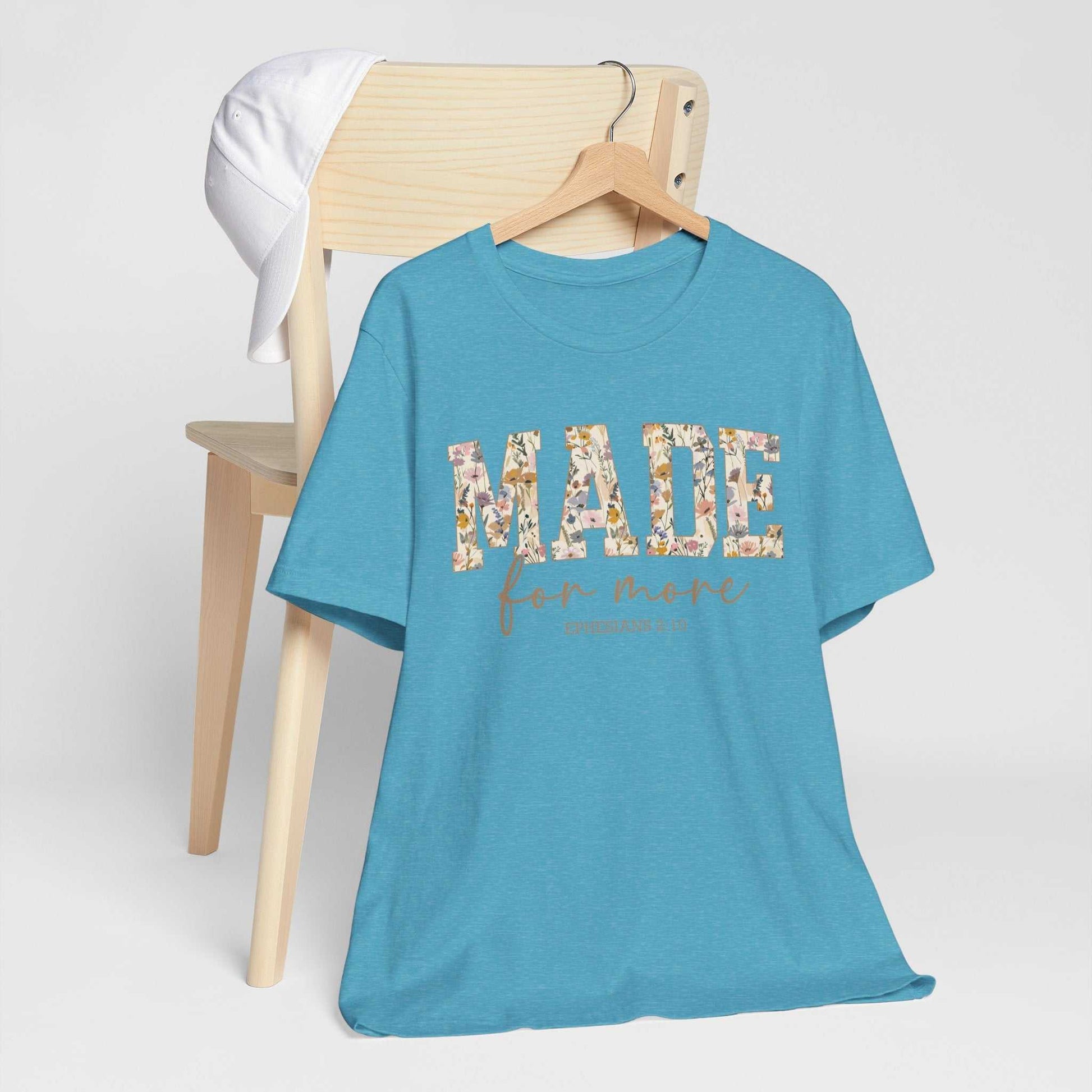 Blue "Made for More" unisex short sleeve tee with floral print text hanging on wooden hanger with white cap on wooden chair in background