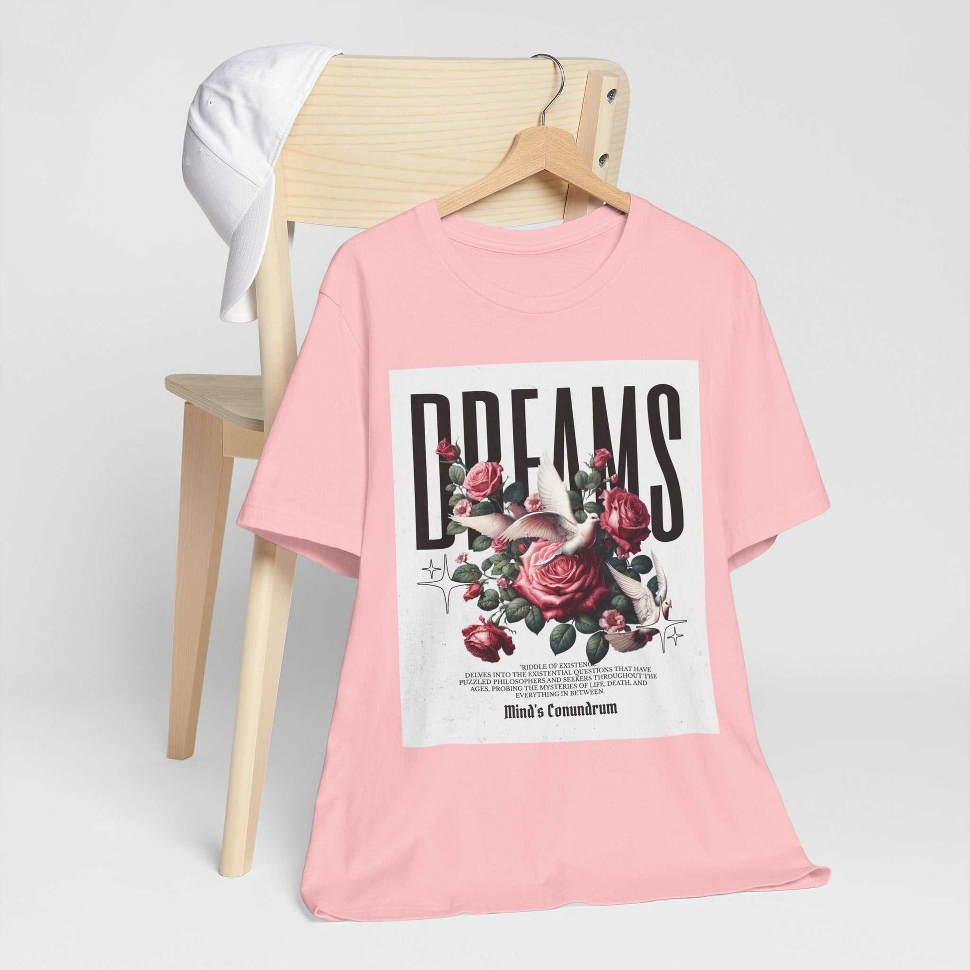 Pink unisex Dreams Floral Tee with rose graphic and bold typography hanging on wooden chair with white cap