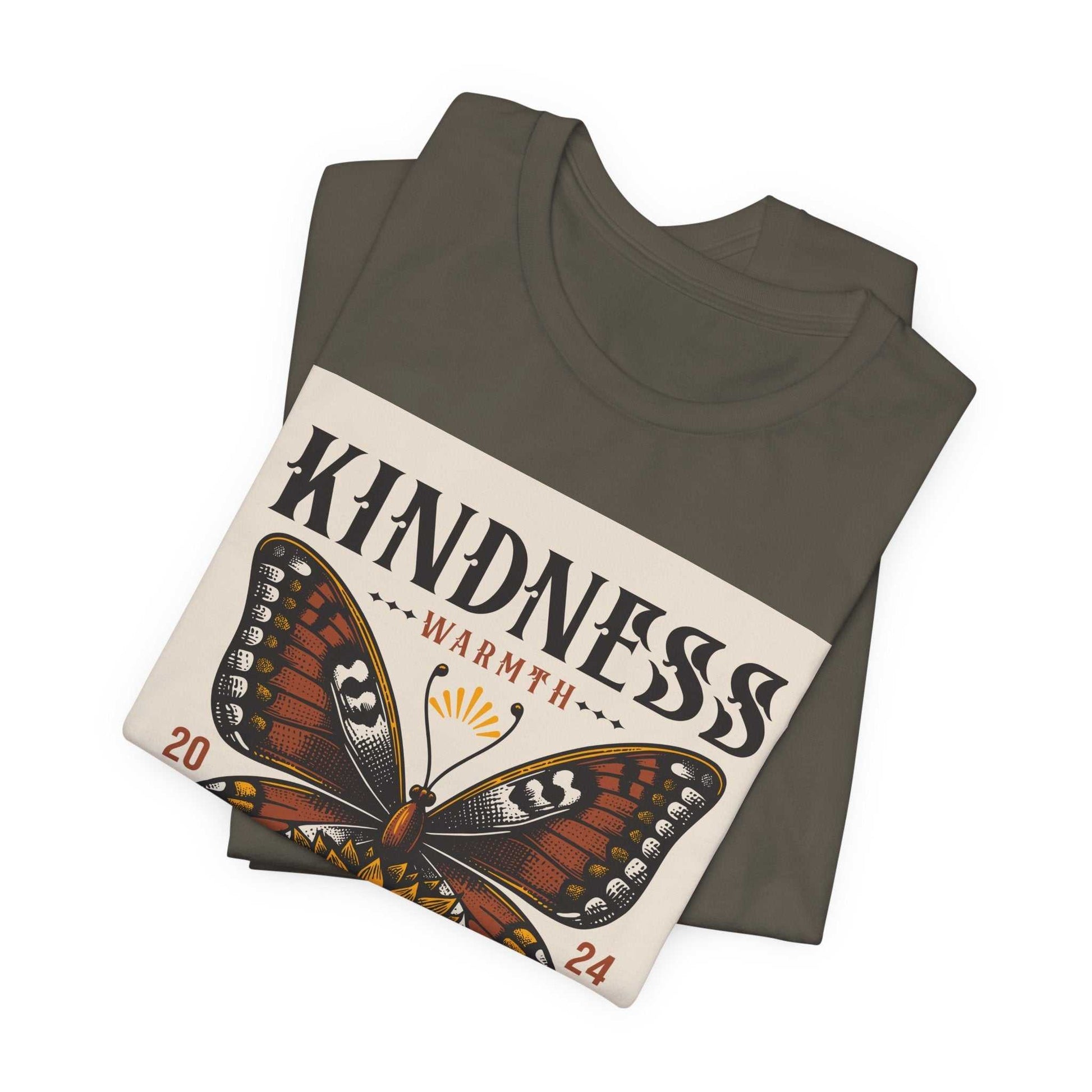 Folded Kindness Butterfly Tee featuring a large, colorful butterfly graphic with the word "KINDNESS" and "WARMTH" printed above and below it on a dark fabric background