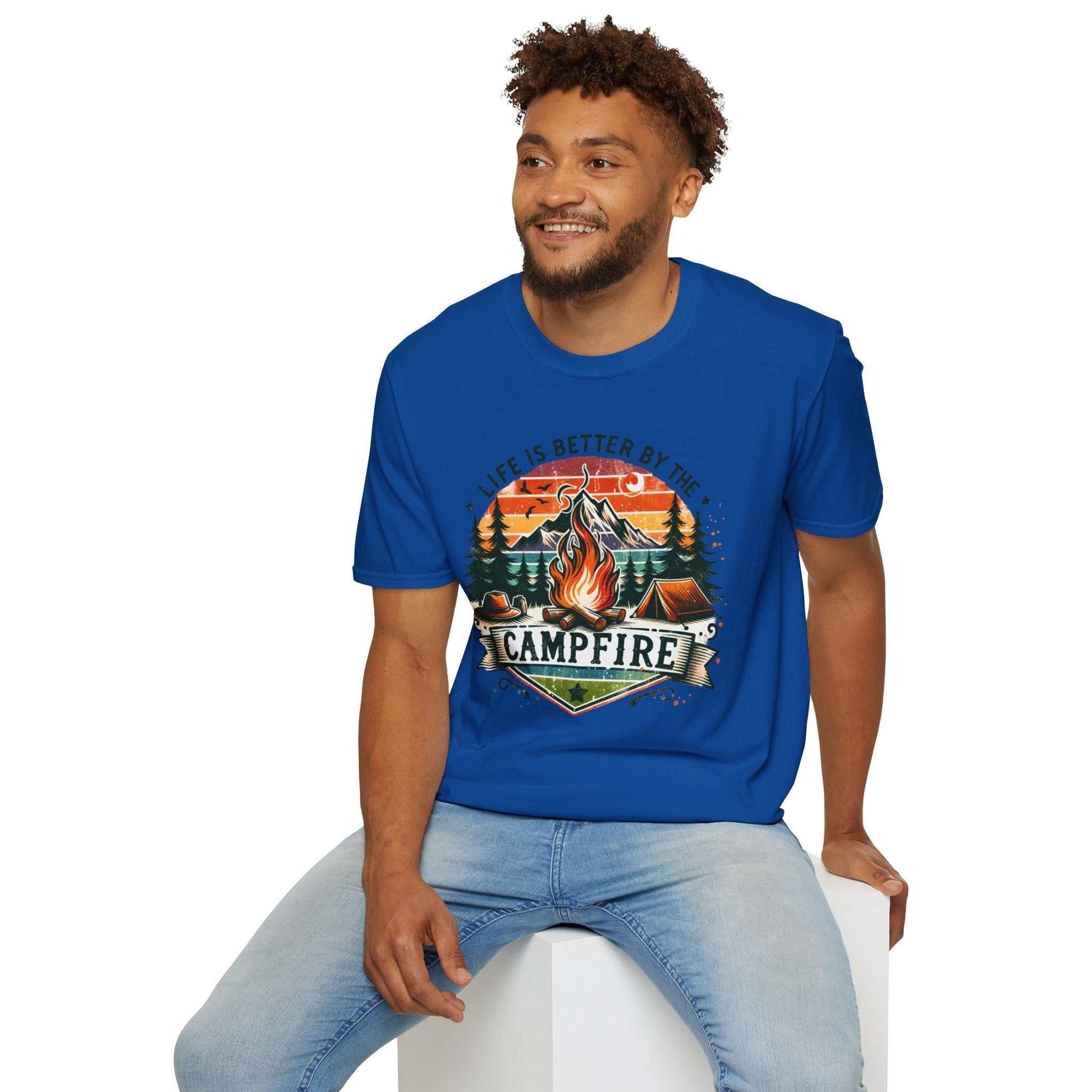 Man wearing blue Camping Adventure unisex t-shirt with campfire and nature design sitting on white block