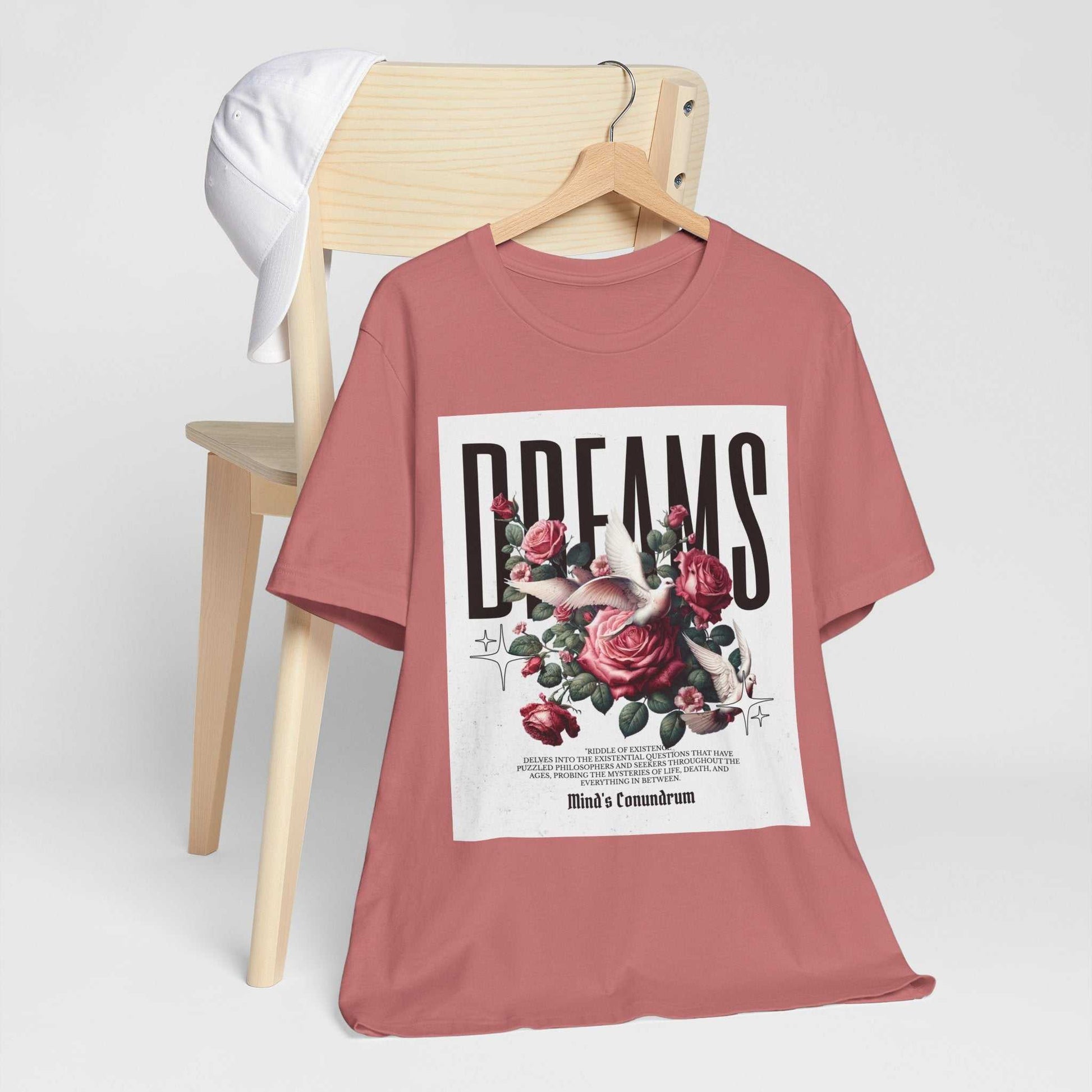 Unisex dusty rose Dreams Floral Tee with bold typography and pink rose graphic on front, displayed on wooden chair with white cap.