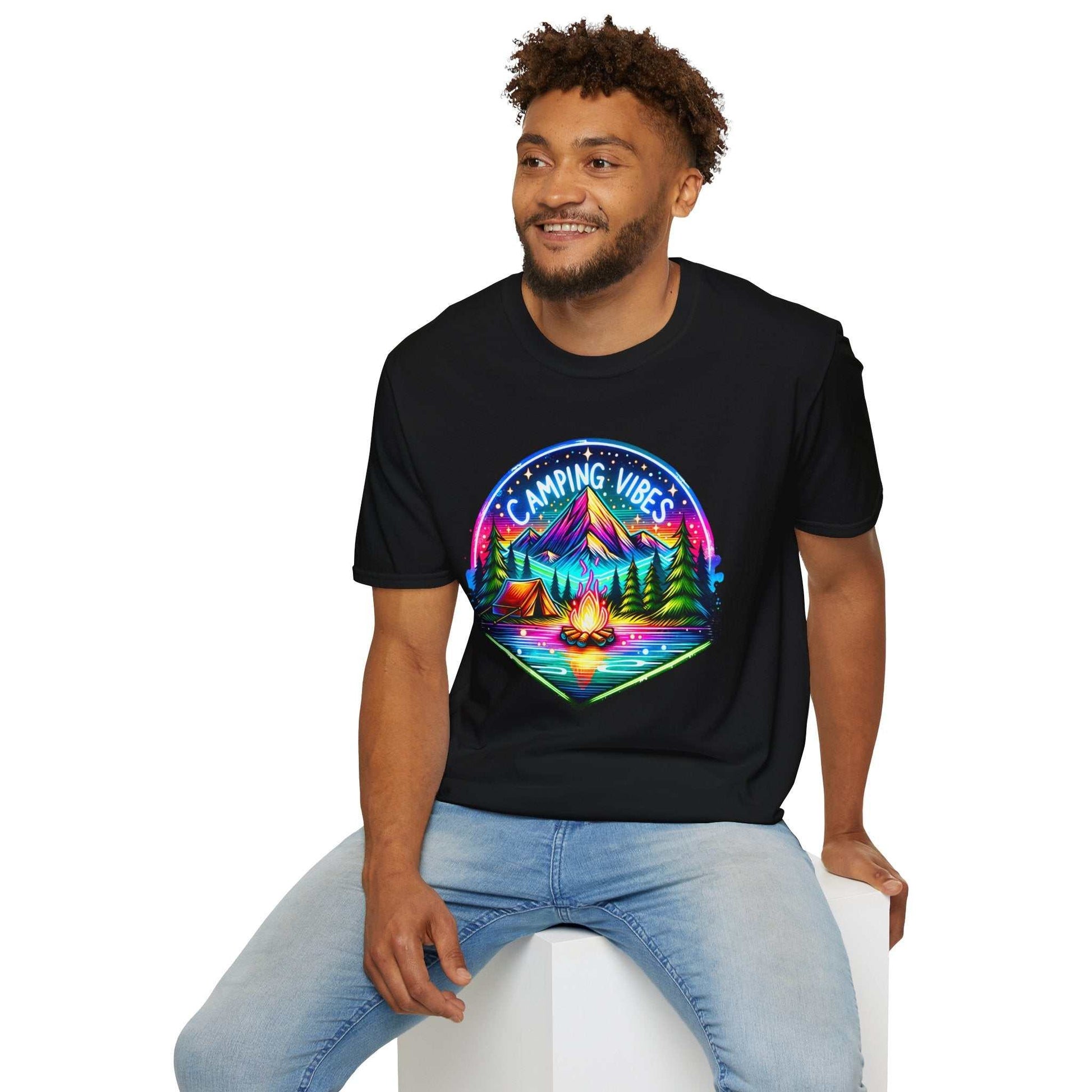 Man wearing black Camping Vibes Unisex Softstyle T-Shirt with colorful mountain, campfire, and forest graphic sitting on white cube