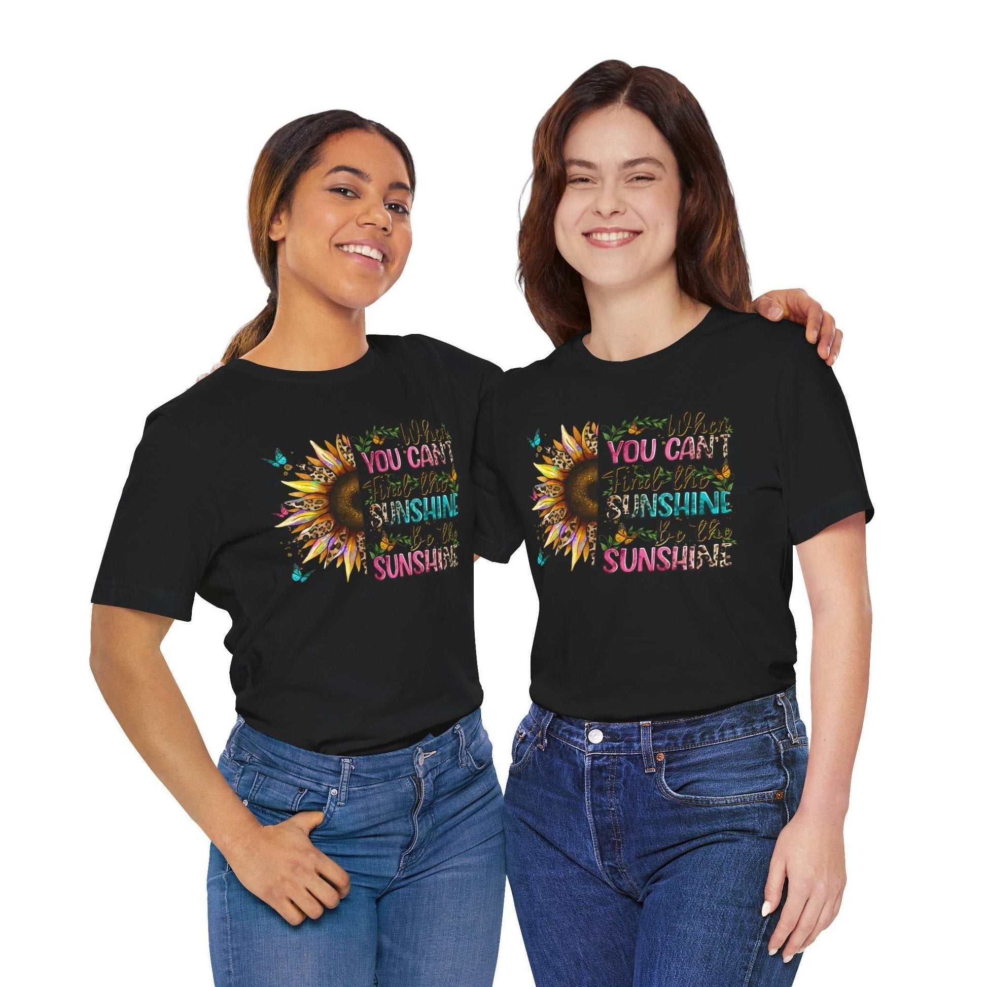 Two women wearing black Sunshine Quote Tee with colorful sunflower and inspirational text design, paired with blue jeans, smiling and posing together