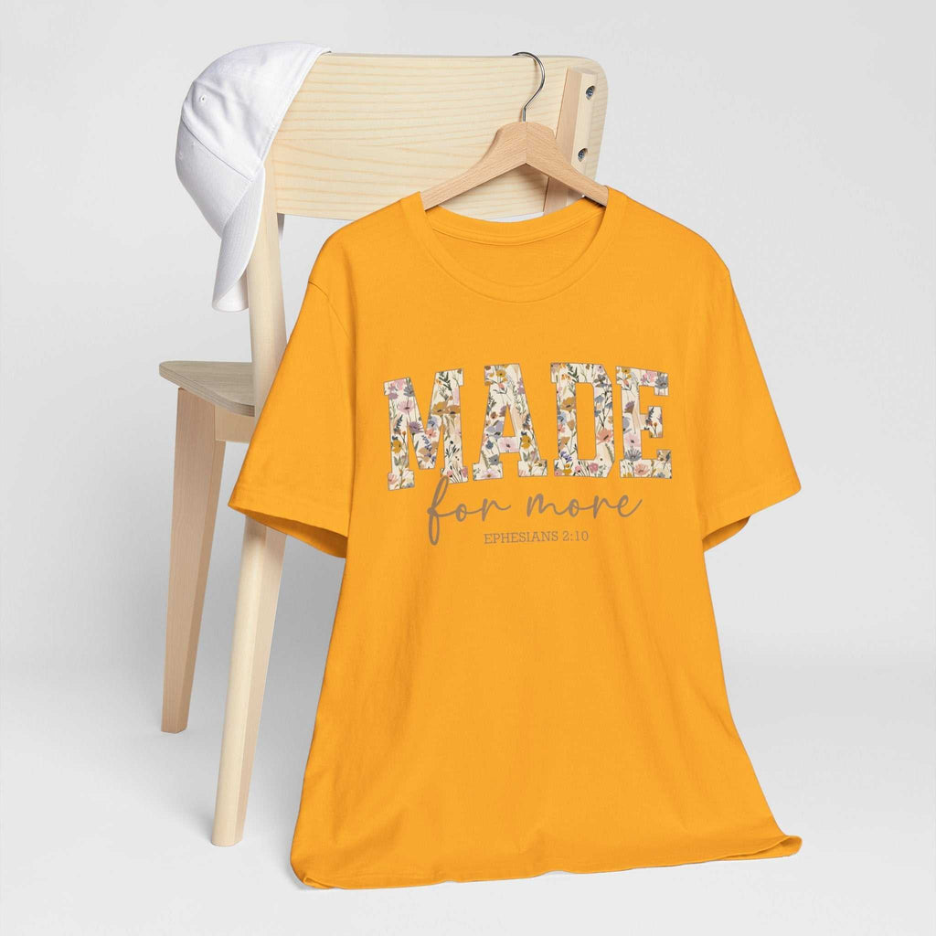 Yellow "Made for More" unisex jersey short sleeve tee with floral text design hanging on wooden hanger by light wood chair with white cap.