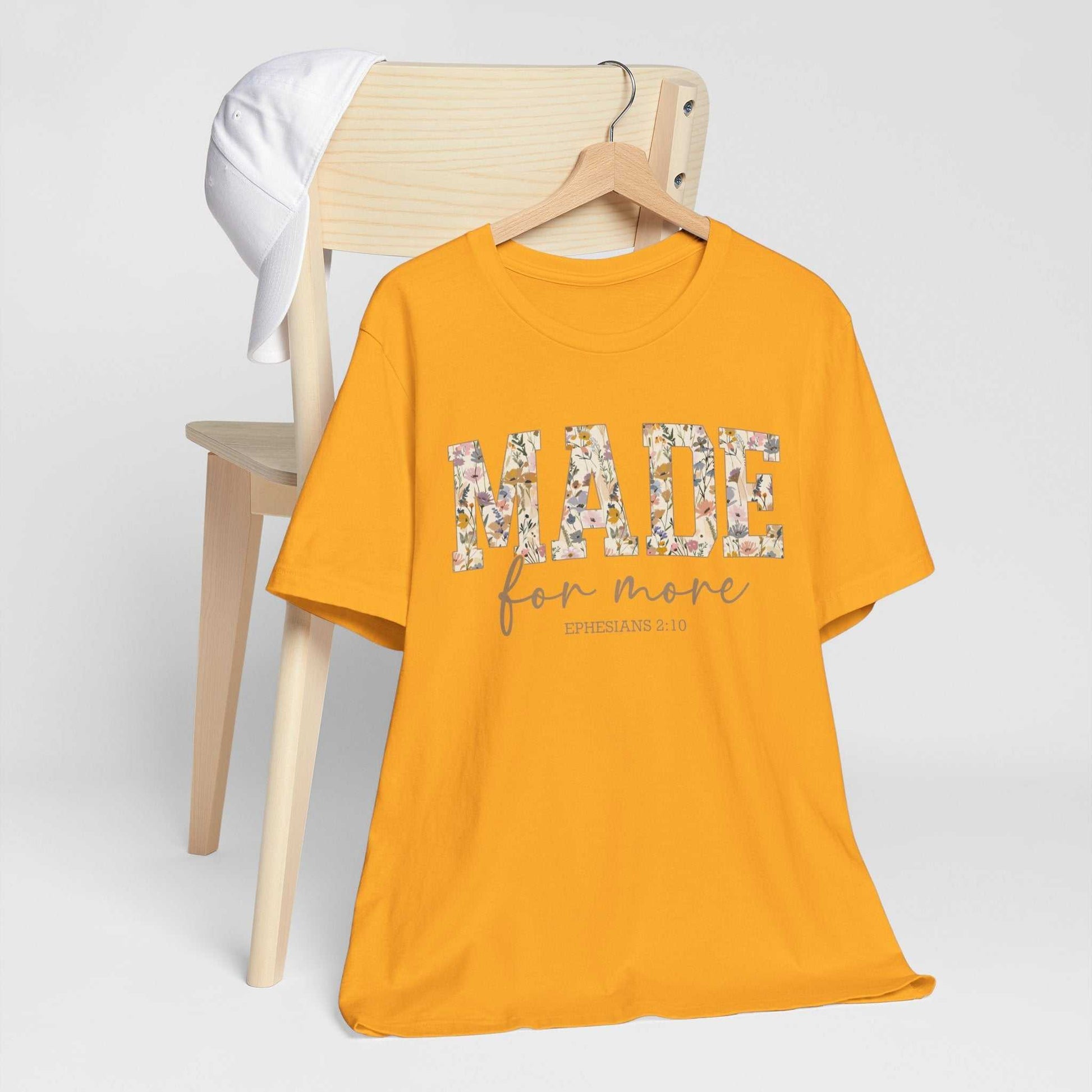 Yellow "Made for More" unisex jersey short sleeve tee with floral text design hanging on wooden hanger by light wood chair with white cap.