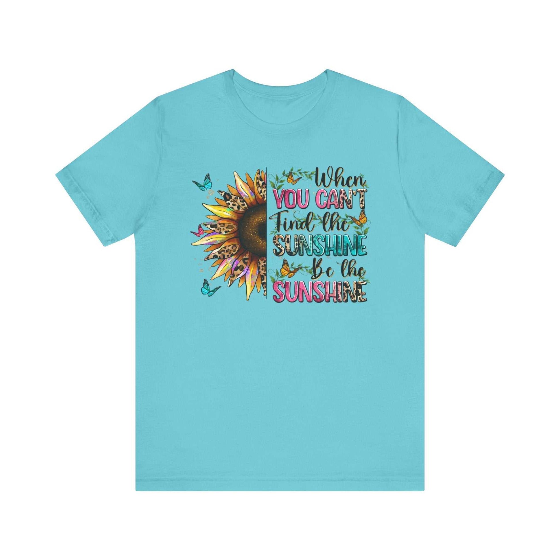 Light blue unisex t-shirt with a colorful sunflower and butterfly design and the quote "When you can't find the sunshine be the sunshine" printed on the front.