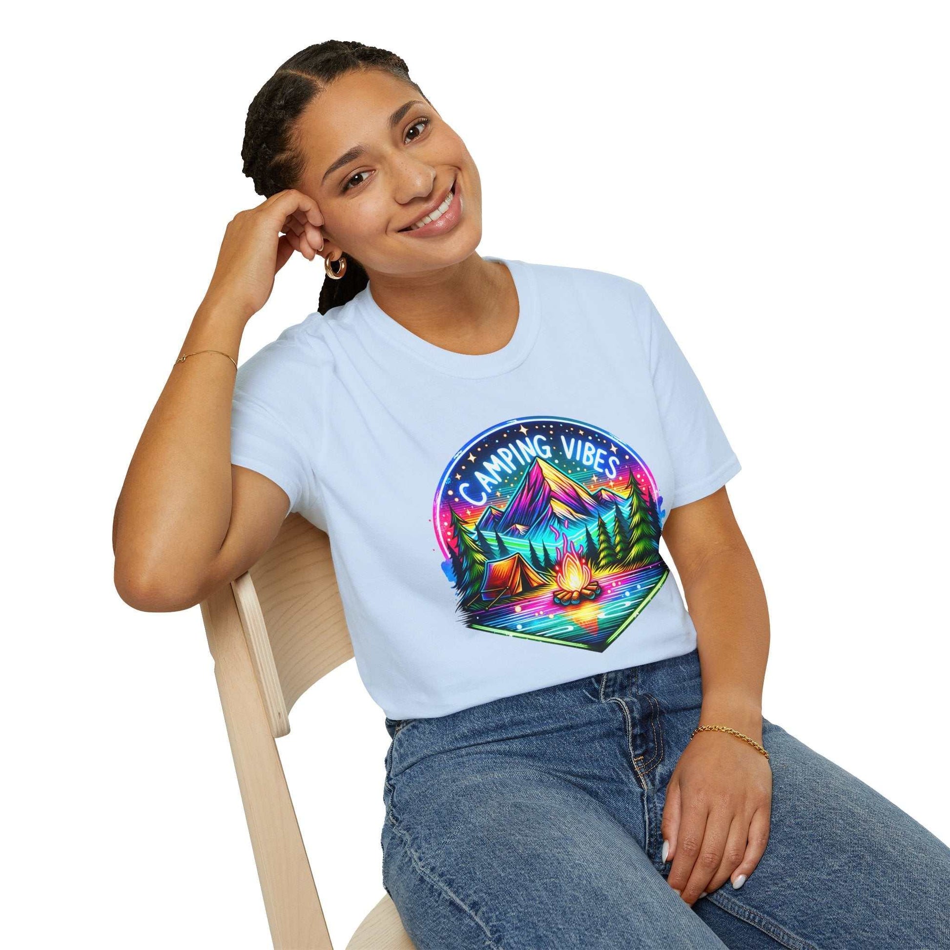 Woman wearing a light blue unisex softstyle t-shirt with a colorful "Camping Vibes" graphic featuring a campfire and mountains sitting on a chair smiling