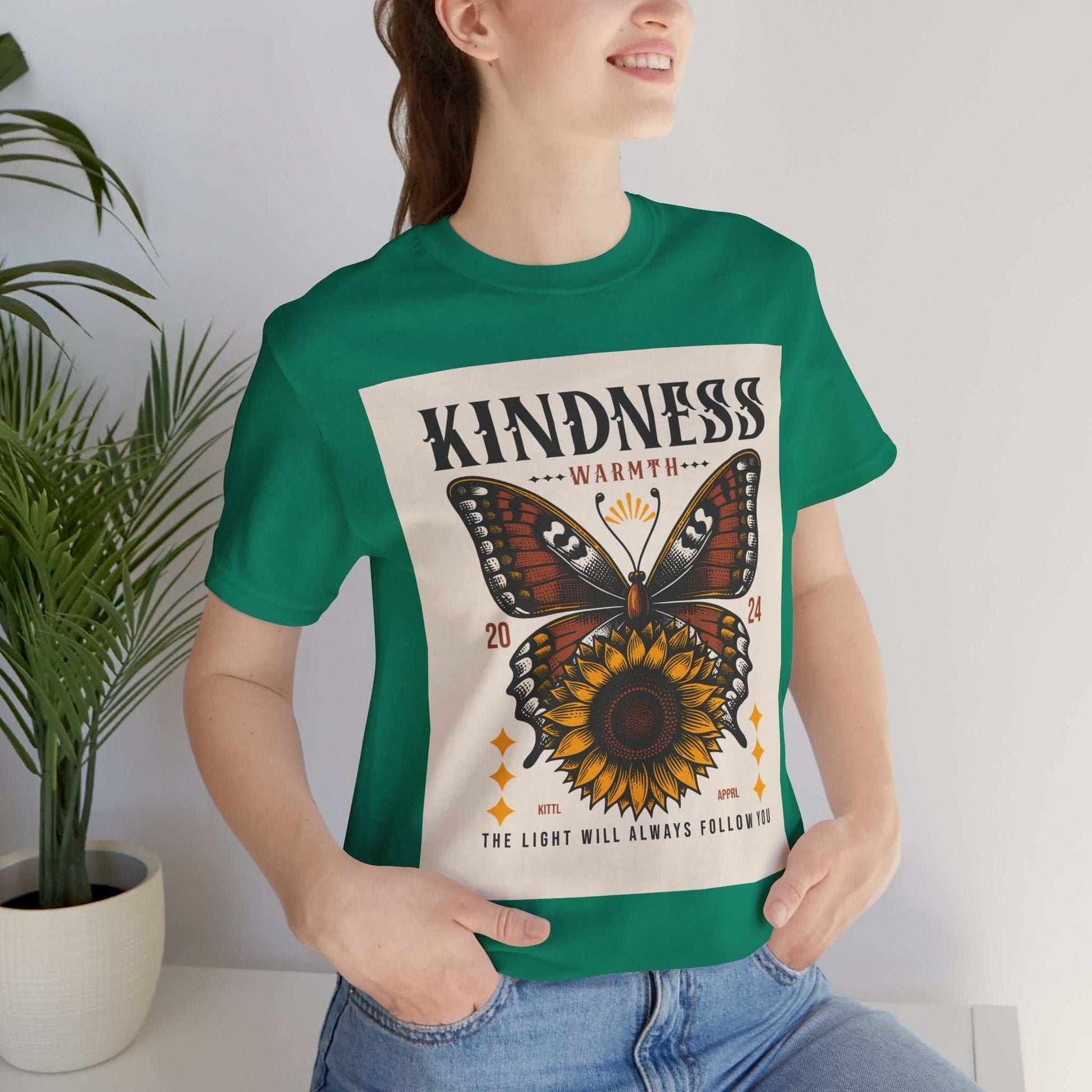 Woman wearing a green Kindness Butterfly Tee with butterfly and sunflower design and text "Kindness Warmth 2024 The light will always follow you"