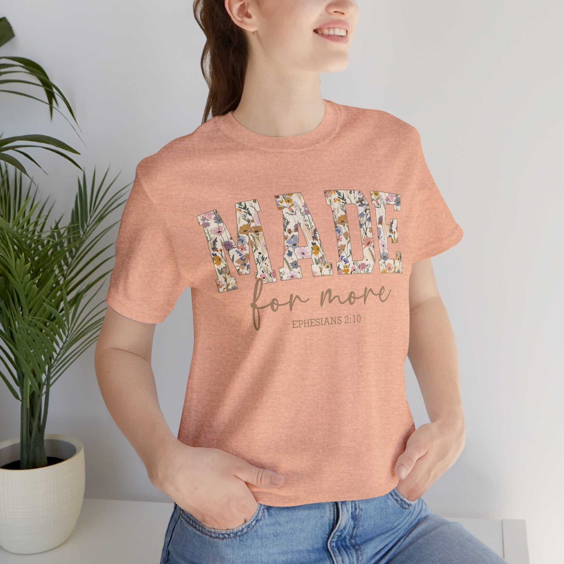 Woman wearing peach Made for More Tee with floral text design and scripture reference, styled casually with blue jeans indoors next to potted plant