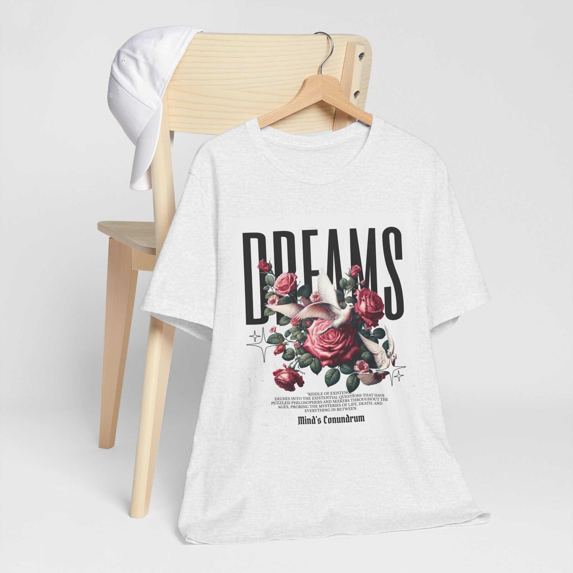 White Dreams Floral Tee with bold typography and pink rose graphic on front, displayed on wooden chair with white cap.
