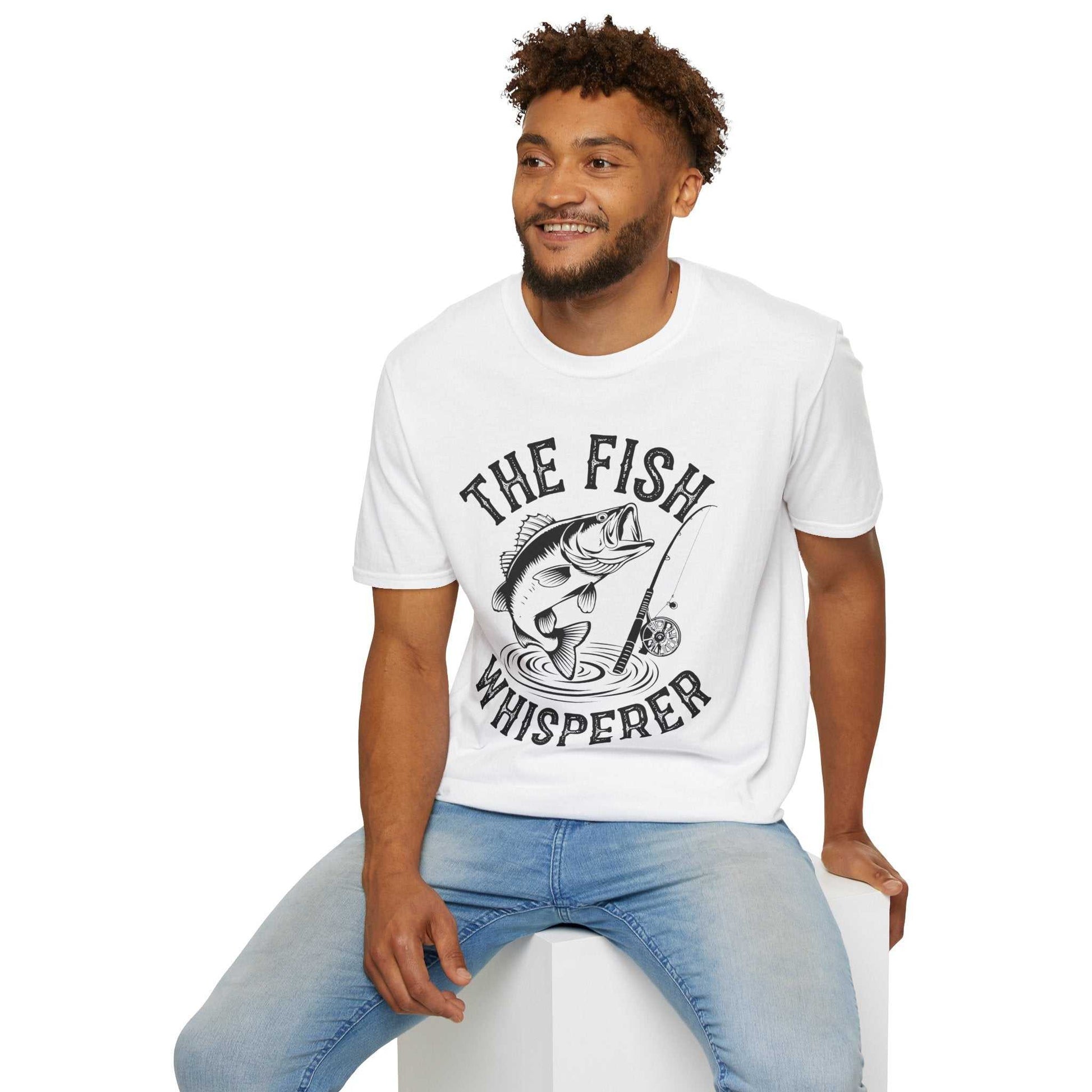 Man wearing white "The Fish Whisperer" graphic unisex softstyle t-shirt and blue jeans sitting on white block