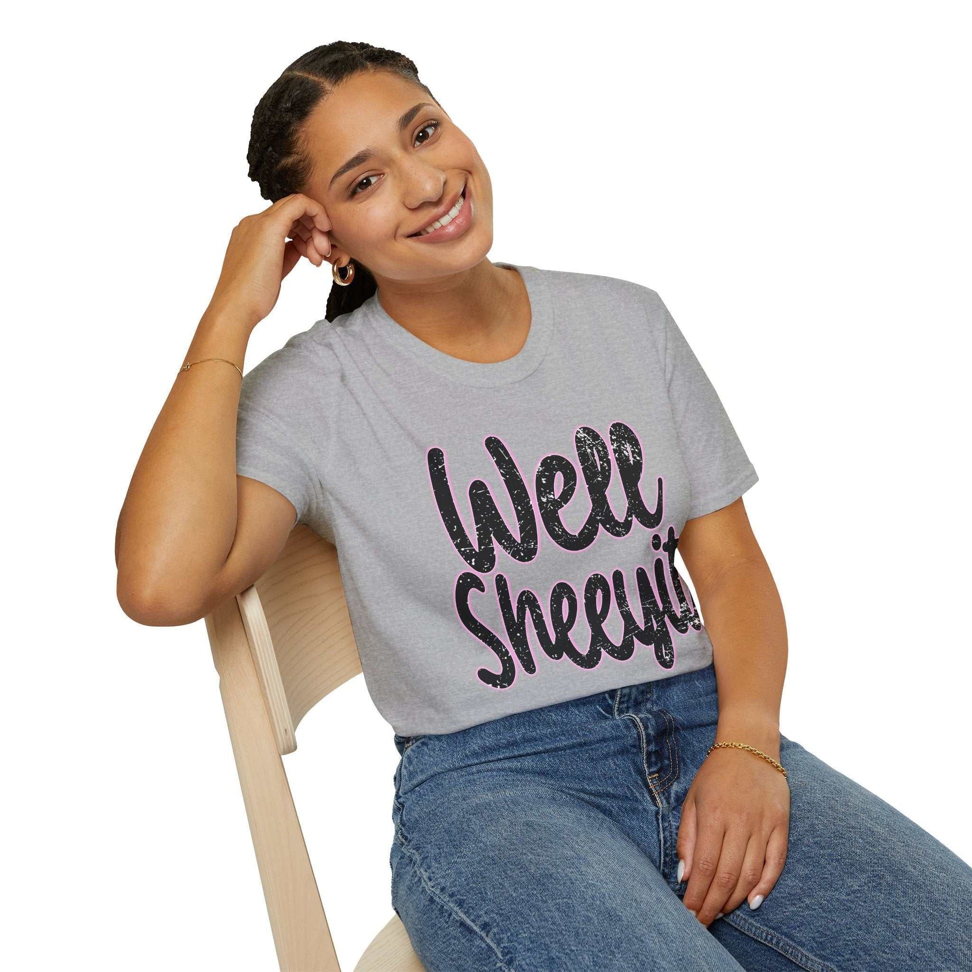 Woman wearing a gray casual statement tee with "Well Sheeyit" printed in large black and pink letters sitting on a wooden chair