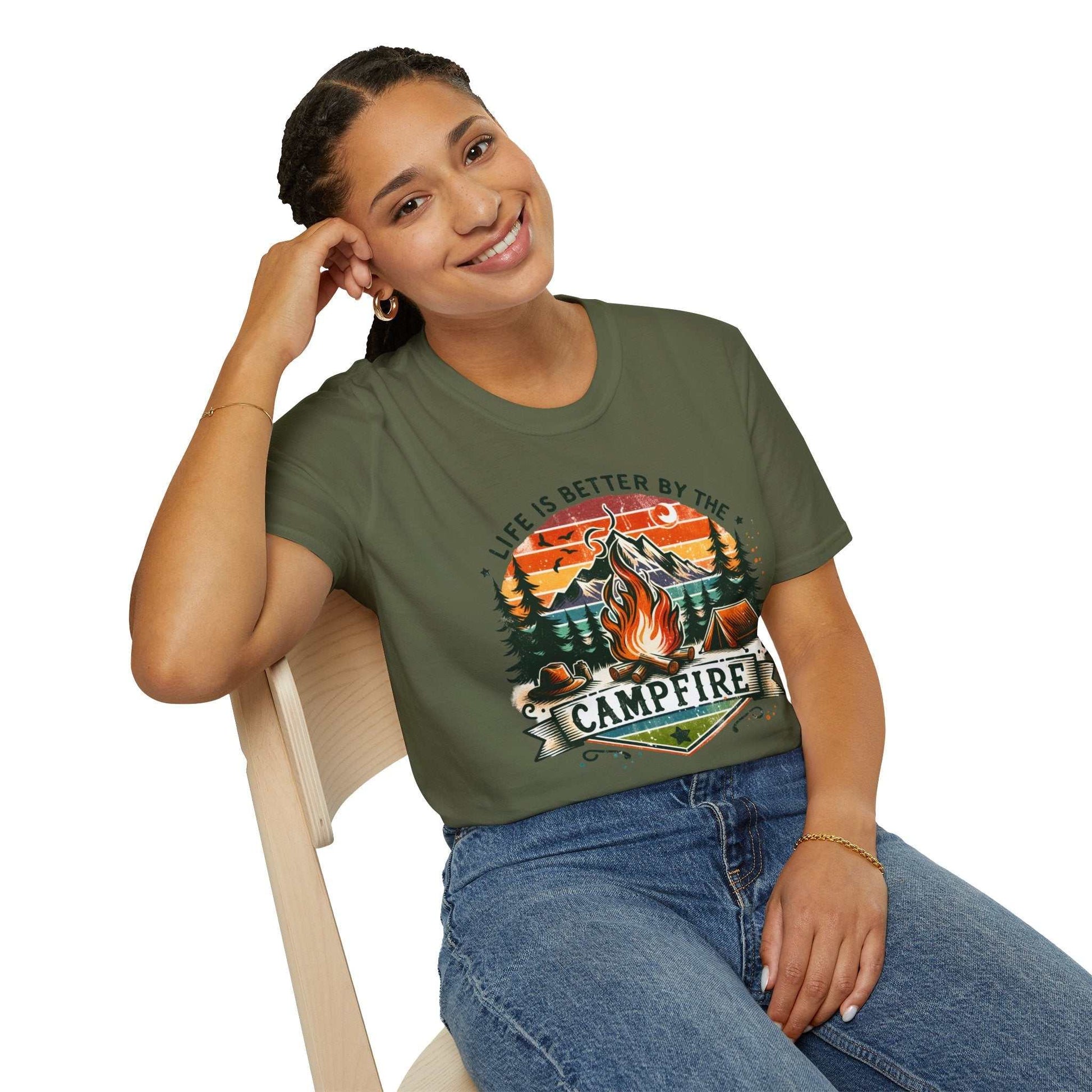 Woman wearing olive green Camping Adventure Unisex T-Shirt with campfire graphic and "Life is Better by the Campfire" text, casual denim jeans seated on chair