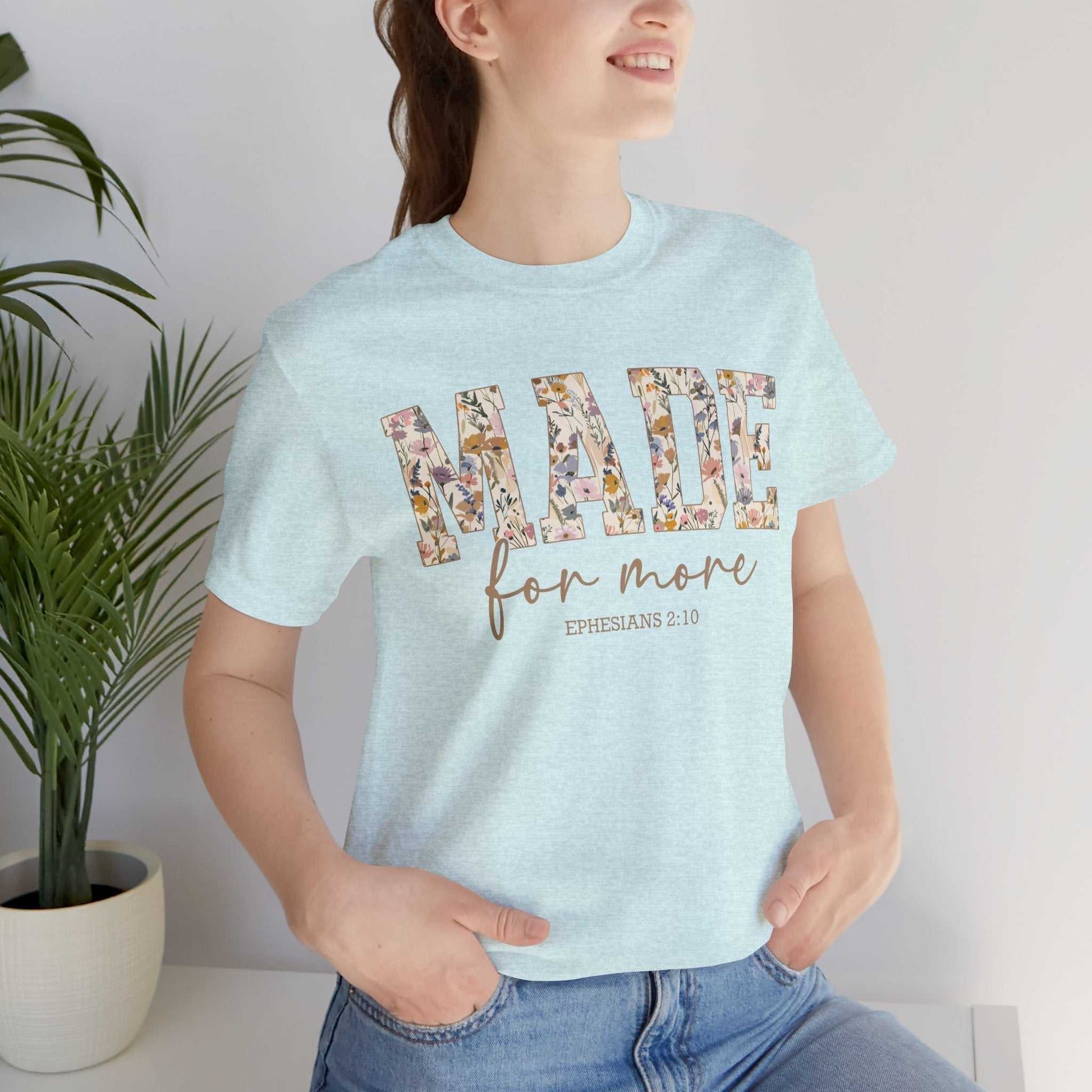 Woman wearing light blue "Made for More" floral text t-shirt with Ephesians 2:10 scripture, styled casual with jeans indoors near plant.
