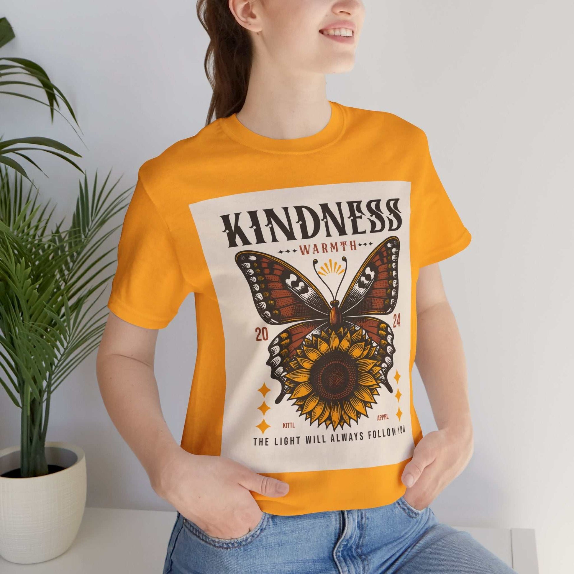 Woman wearing orange Kindness Butterfly Tee with butterfly and sunflower graphic and text "Kindness Warmth The Light Will Always Follow"