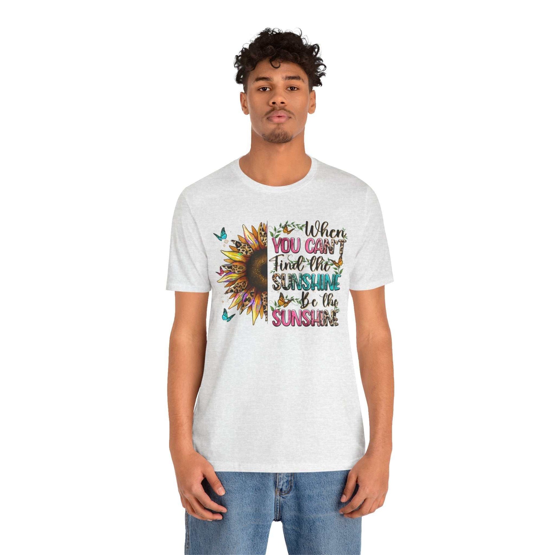 Man wearing white Sunshine Quote Tee with colorful sunflower and uplifting text design, paired with blue jeans.