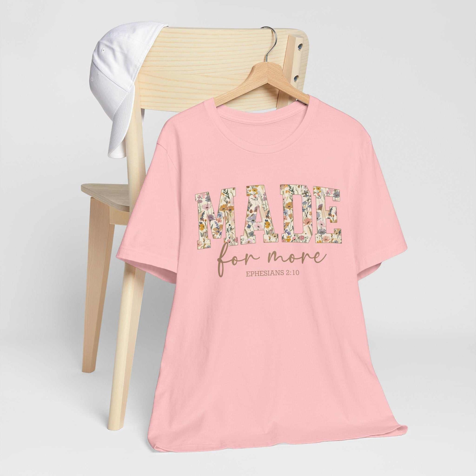 Pink Made for More Tee with floral print text hanging on wooden hanger on chair with white cap nearby