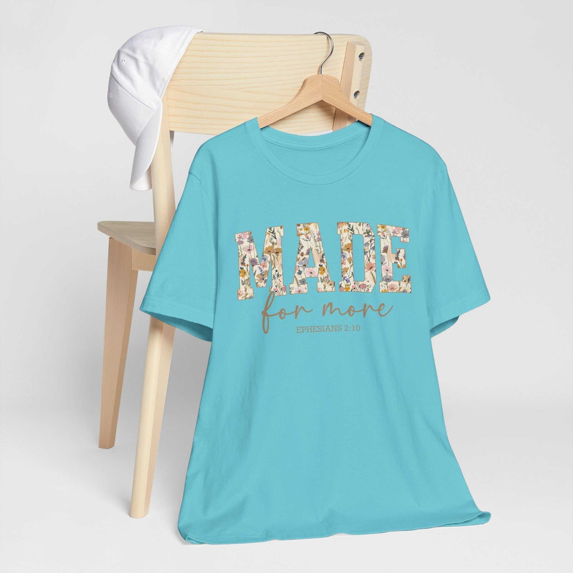 Light blue Made for More Tee with floral patterned lettering, hanging on a wooden hanger on a chair with a white cap resting on the chair back