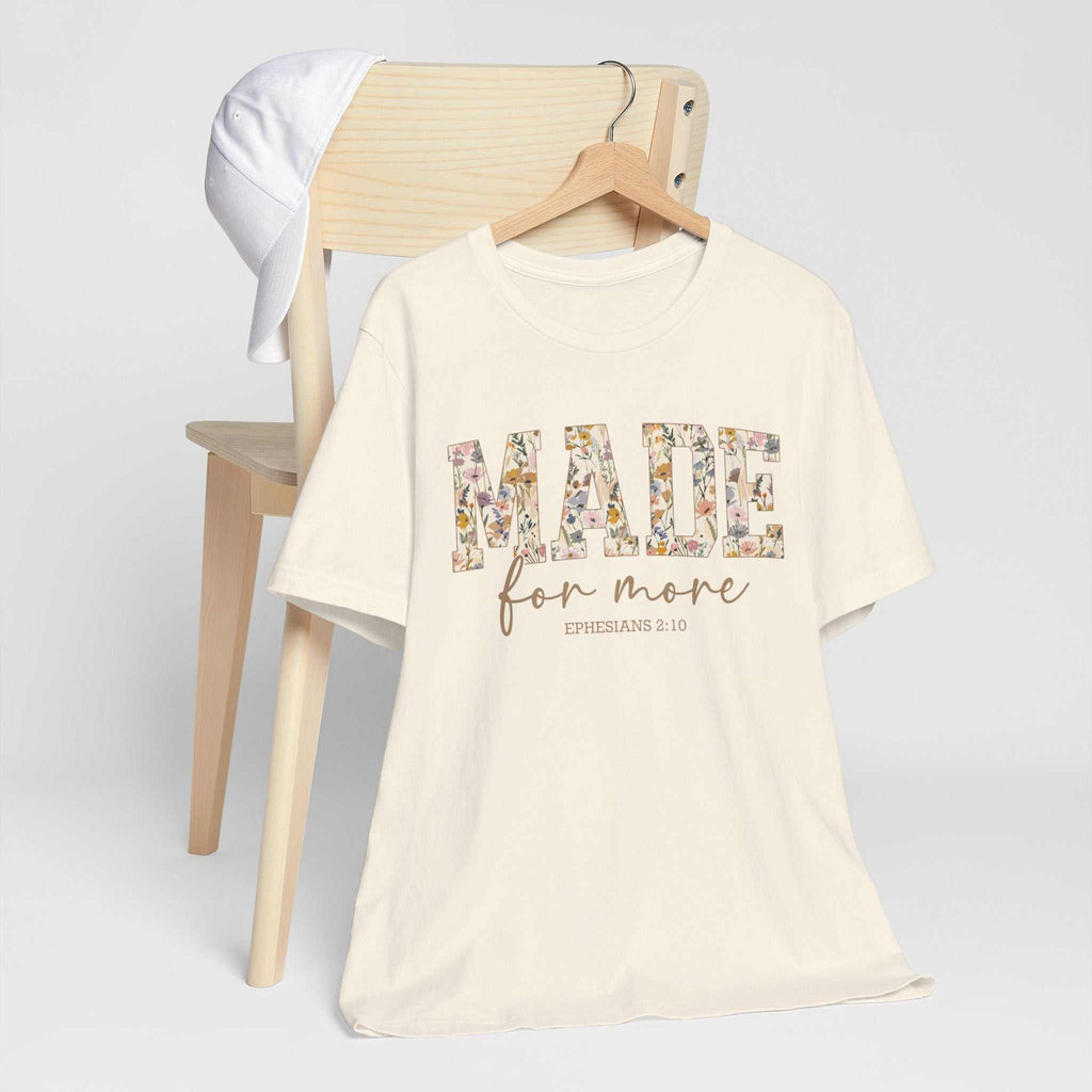 Cream-colored Made for More Tee with floral patterned text and Ephesians 2:10 scripture, displayed on a wooden chair with a white cap hanging on it.