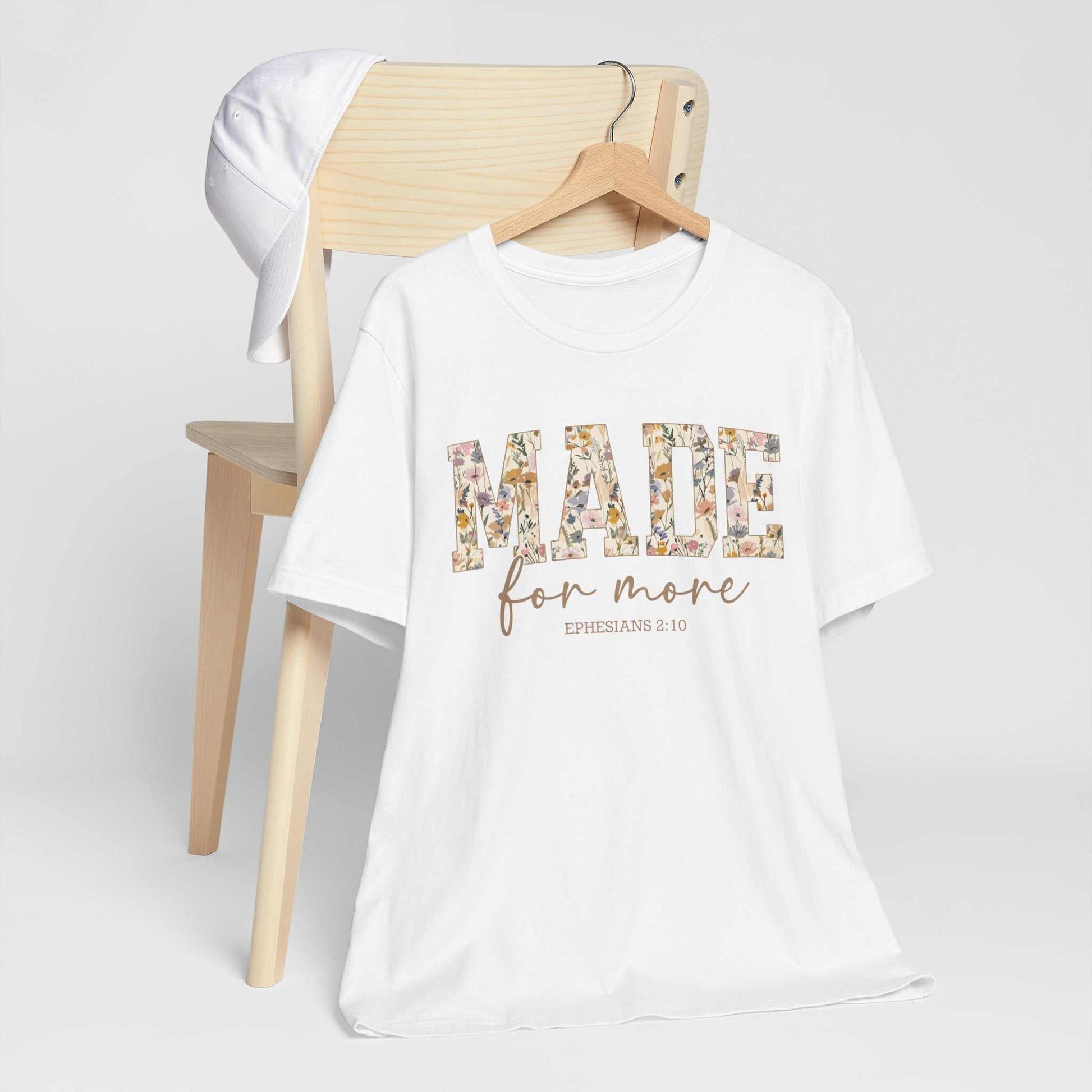 White "Made for More Tee" with floral text design, displayed on a wooden chair with a white cap hanging on the chair back, unisex short sleeve cotton t-shirt.