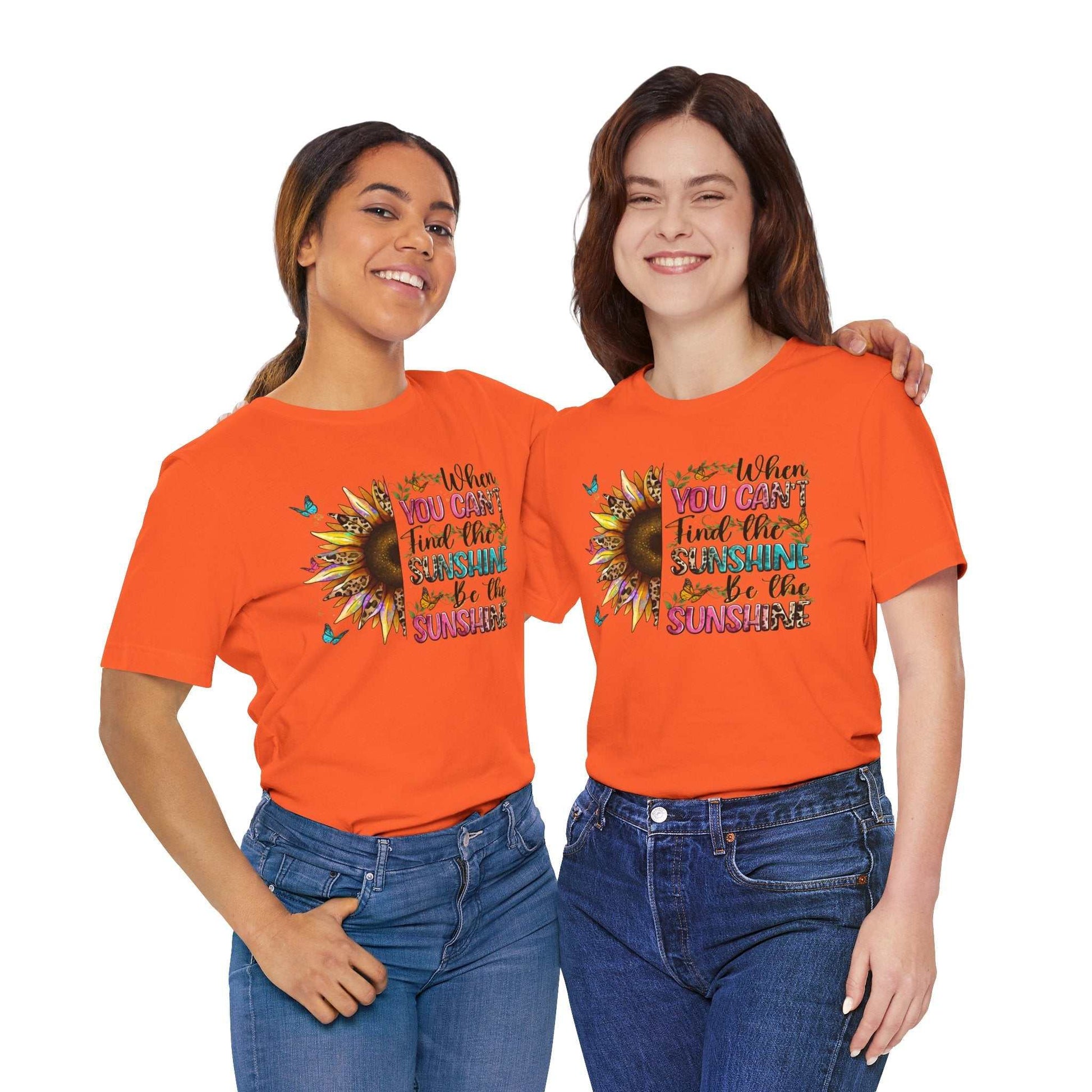 Two smiling women wearing bright orange Sunshine Quote Tee shirts with a colorful sunflower and motivational text design, styled casually with jeans.