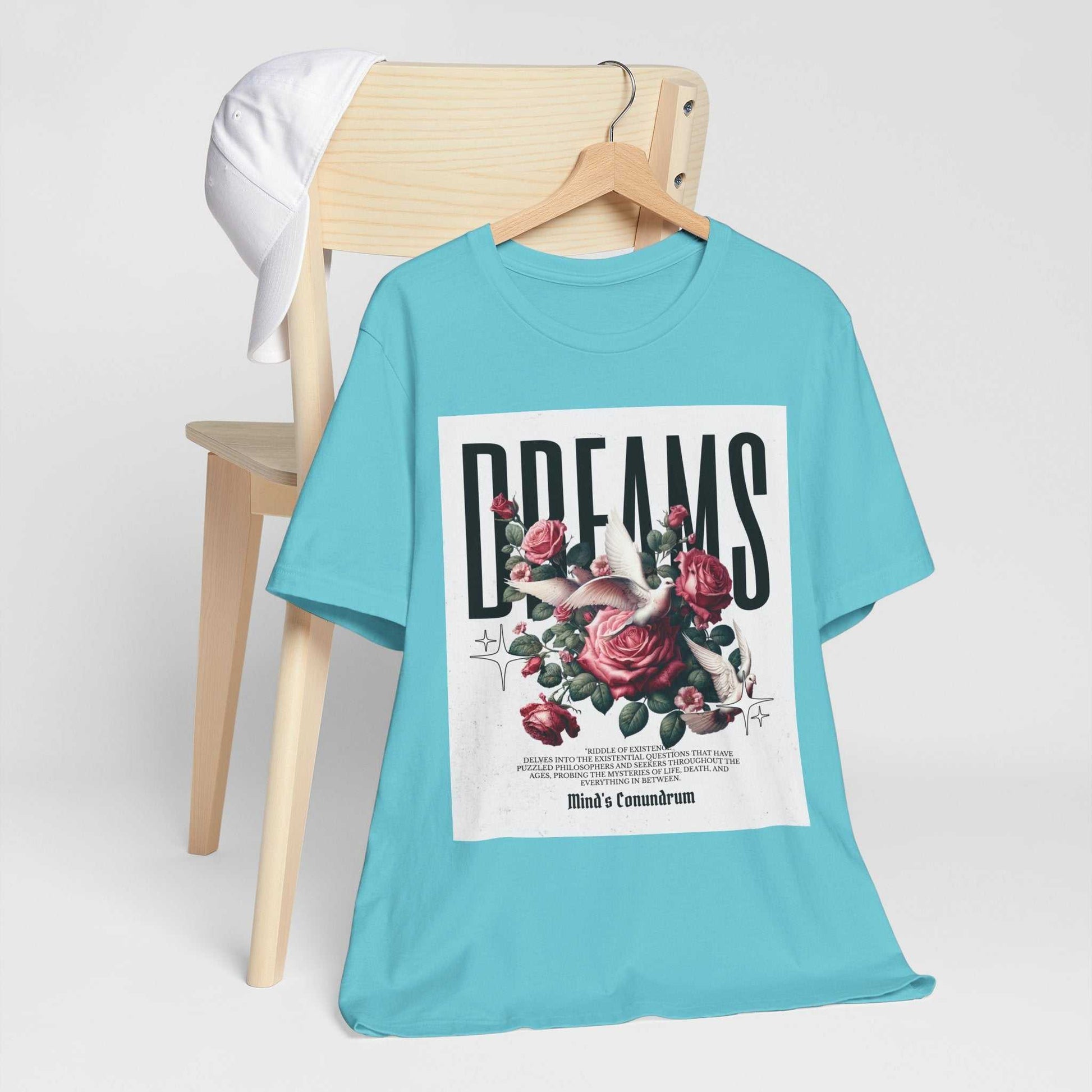 Light blue Dreams Floral Tee with bold black text and pink rose graphic on front, displayed on wooden chair with white cap.