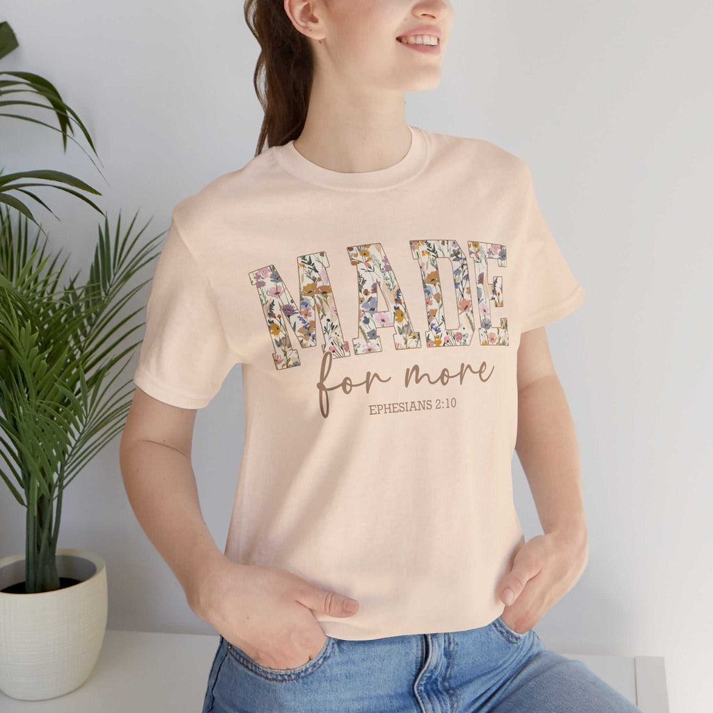 Woman wearing beige Made for More Tee with floral print text and scripture reference Ephesians 2:10, styled casually with jeans indoors near plant