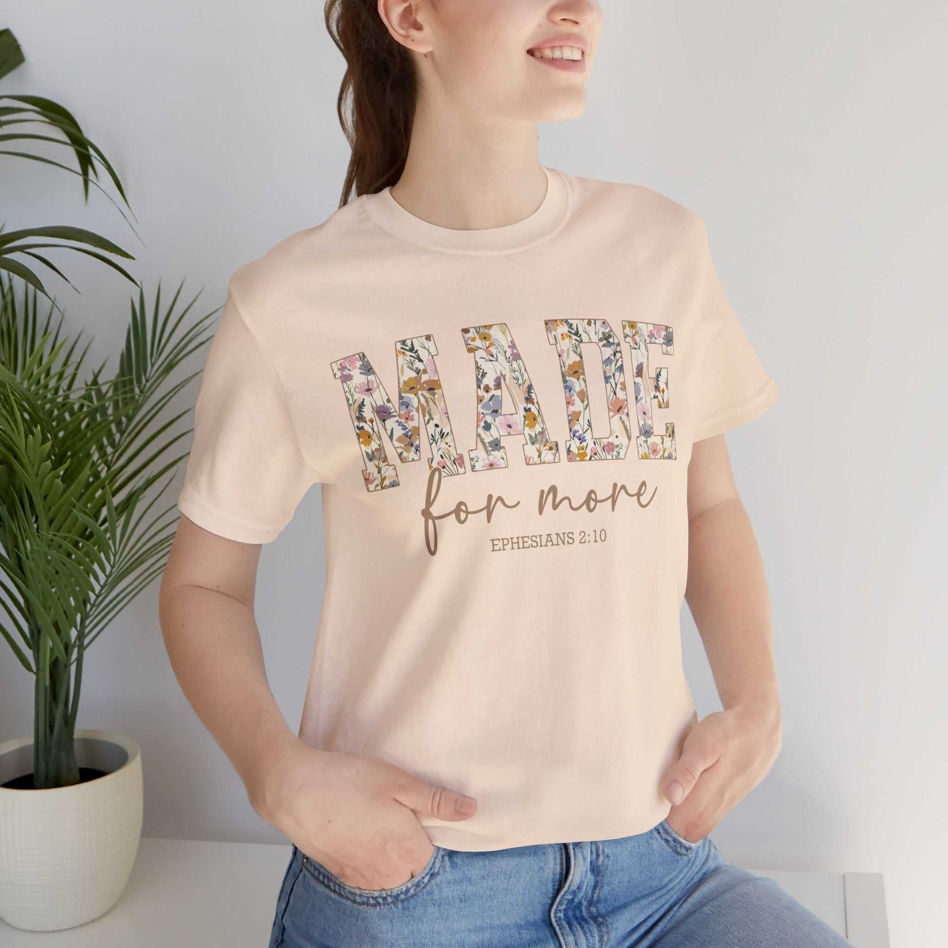 Woman wearing beige Made for More Tee with floral print text and scripture reference Ephesians 2:10, styled casually with jeans indoors near plant