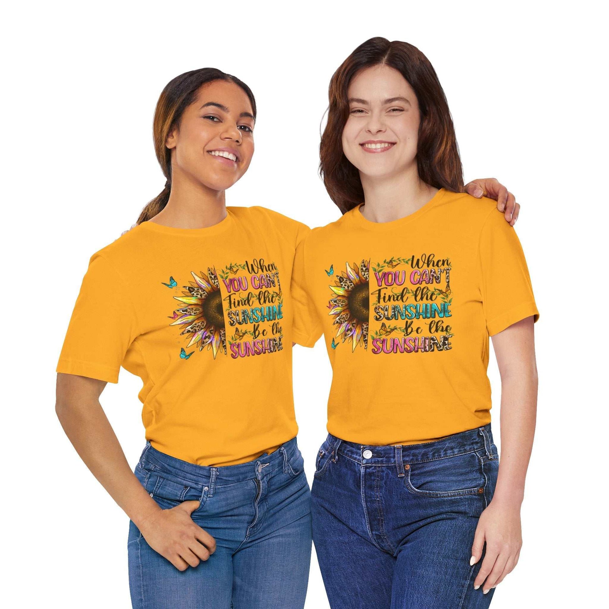 Two women wearing bright yellow Sunshine Quote Tees with a colorful sunflower and motivational text design, paired with casual blue jeans.