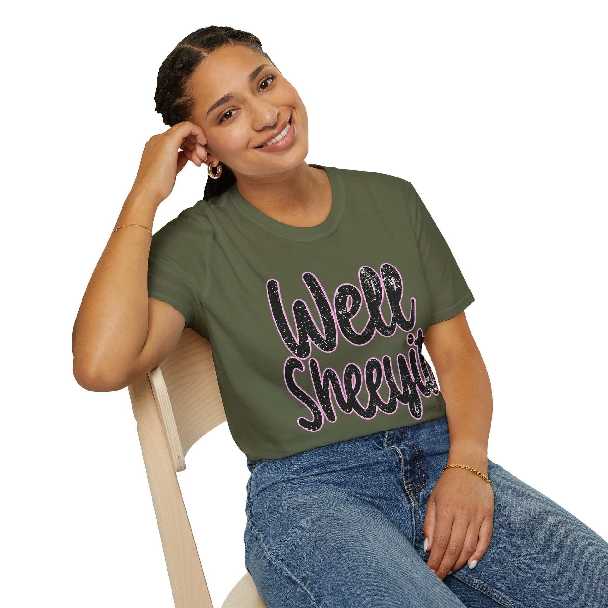 Woman wearing olive green casual statement tee with funny "Well Sheeyit" text, paired with blue jeans, sitting on wooden chair