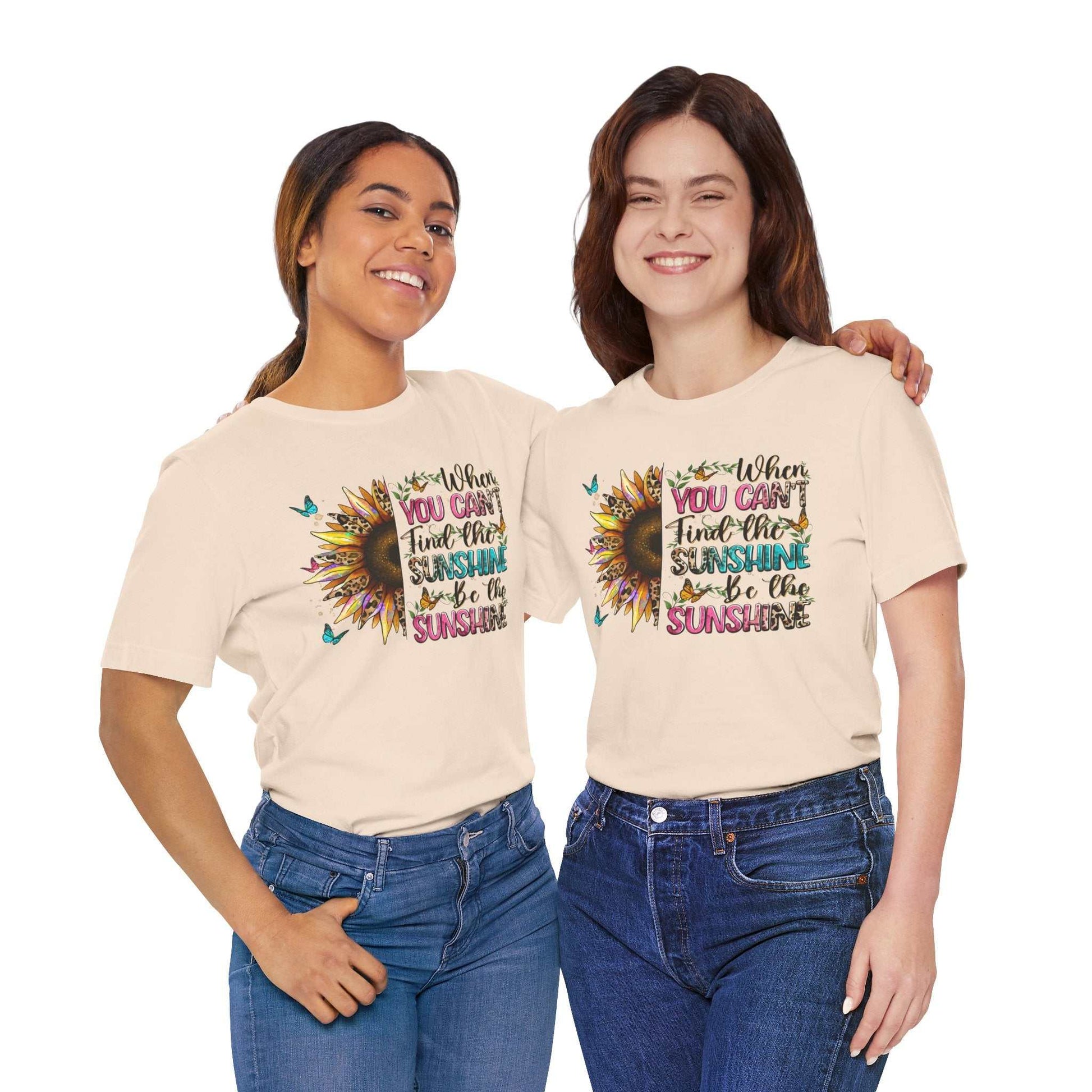 Two women wearing beige Sunshine Quote Tees with a colorful sunflower and motivational text design, styled with casual blue jeans.