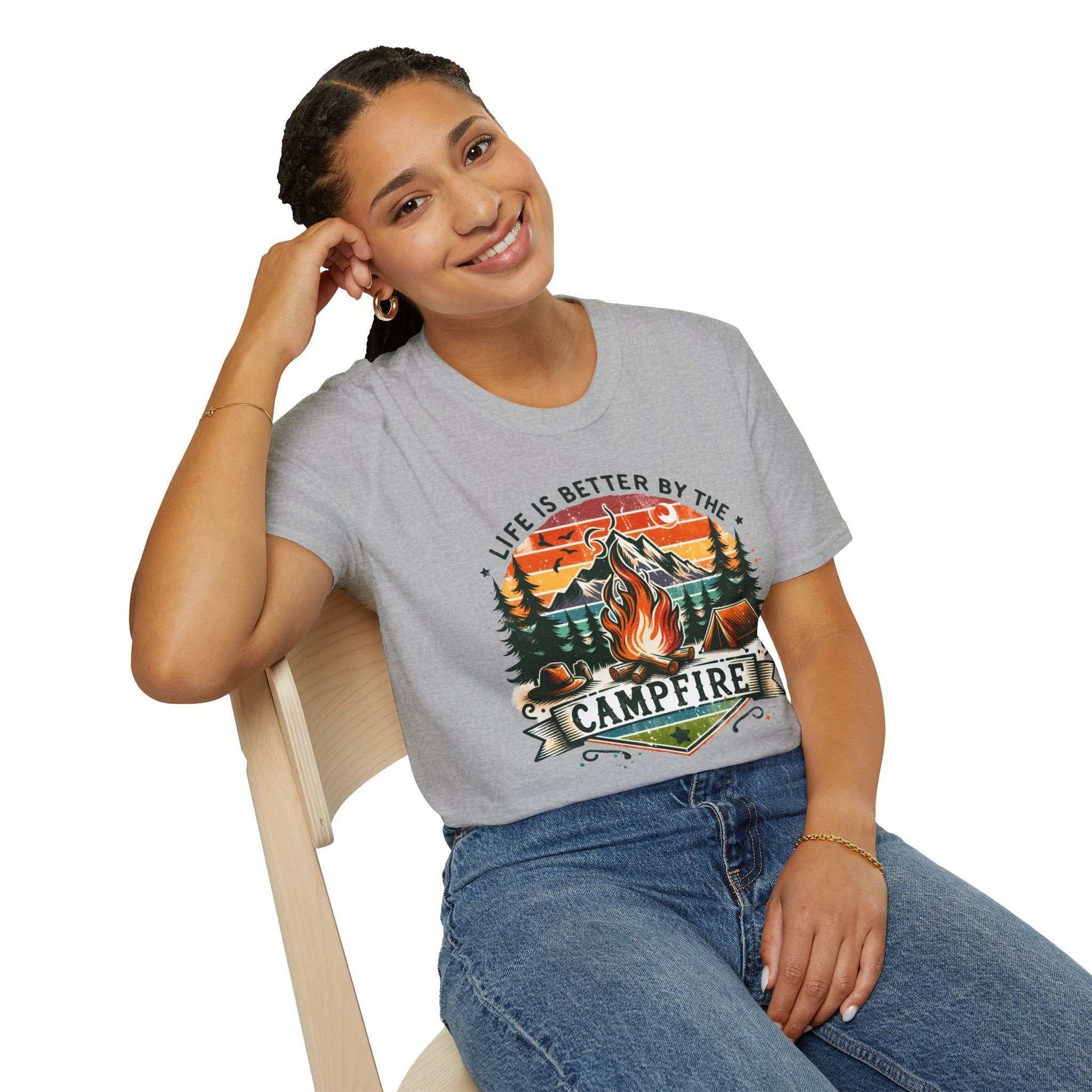 Woman wearing grey Camping Adventure Unisex T-Shirt with campfire graphic and "Life is Better by the Campfire" text, sitting on chair.