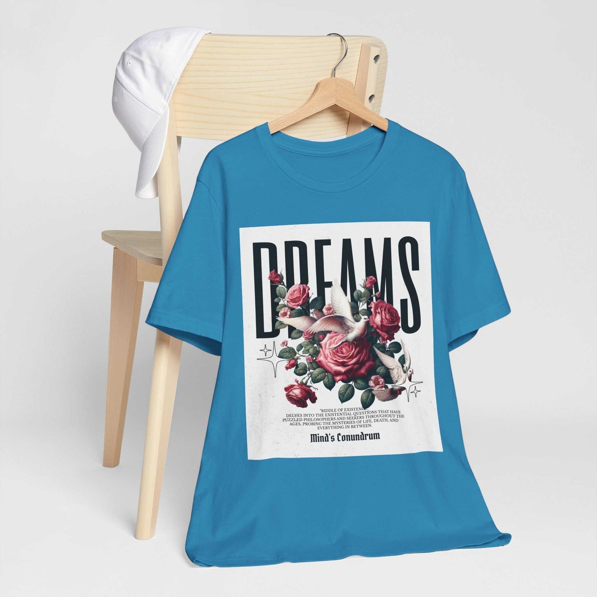 Blue Dreams Floral Tee with rose graphic and bold typography hanging on wooden chair with white cap.