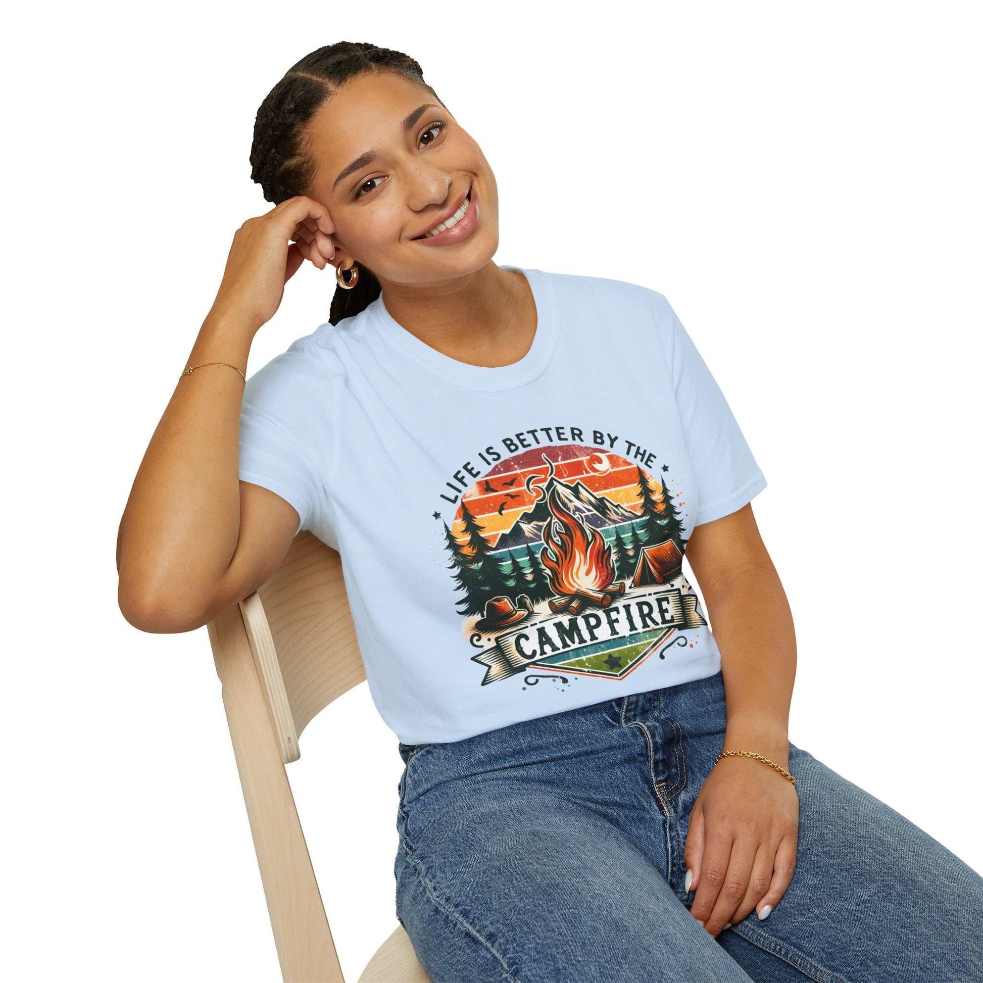 Woman wearing a light blue Camping Adventure Unisex T-Shirt with "Life is Better by the Campfire" design sitting on a wooden chair
