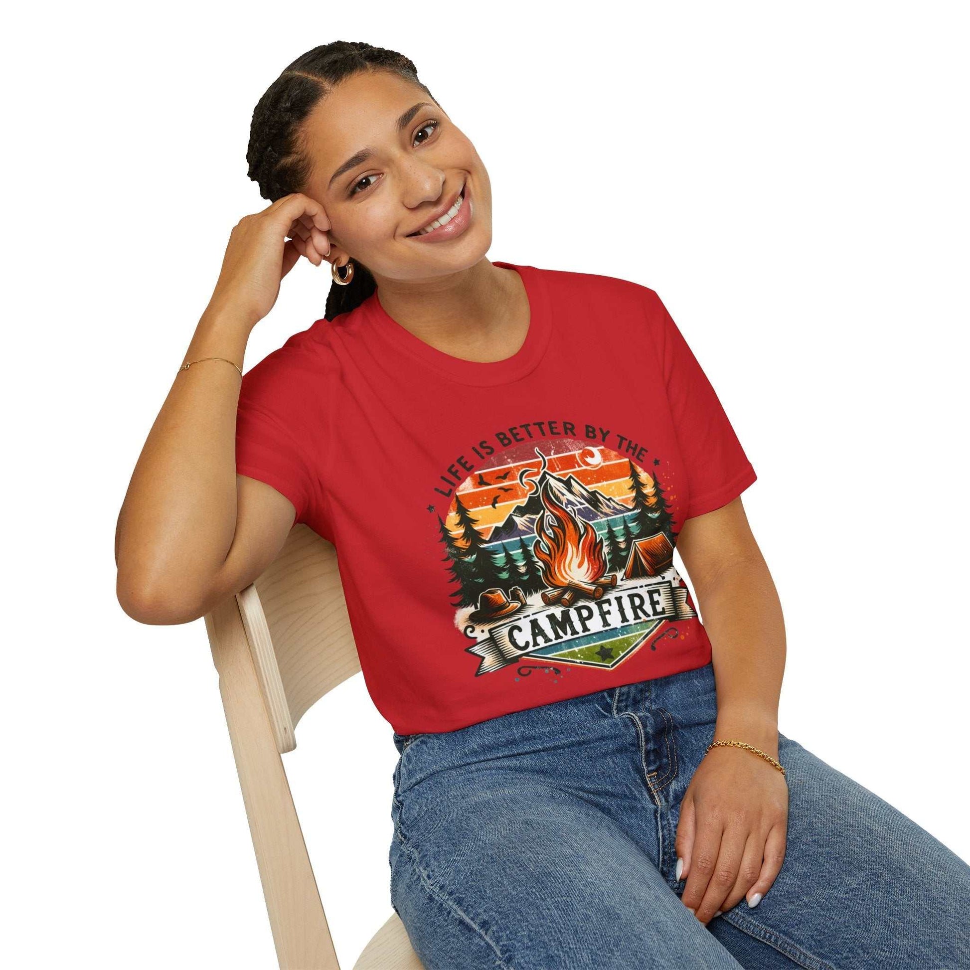 Woman wearing a red Camping Adventure Unisex T-Shirt with "Life is Better by the Campfire" graphic sitting on a wooden chair