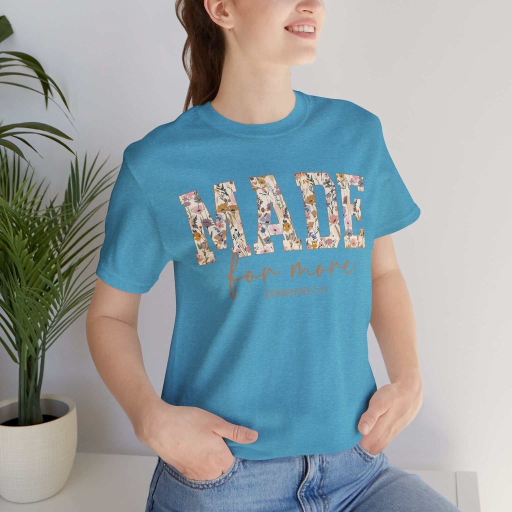 Woman wearing a blue "Made for More Tee" with floral patterned text and casual jeans standing indoors near a plant.