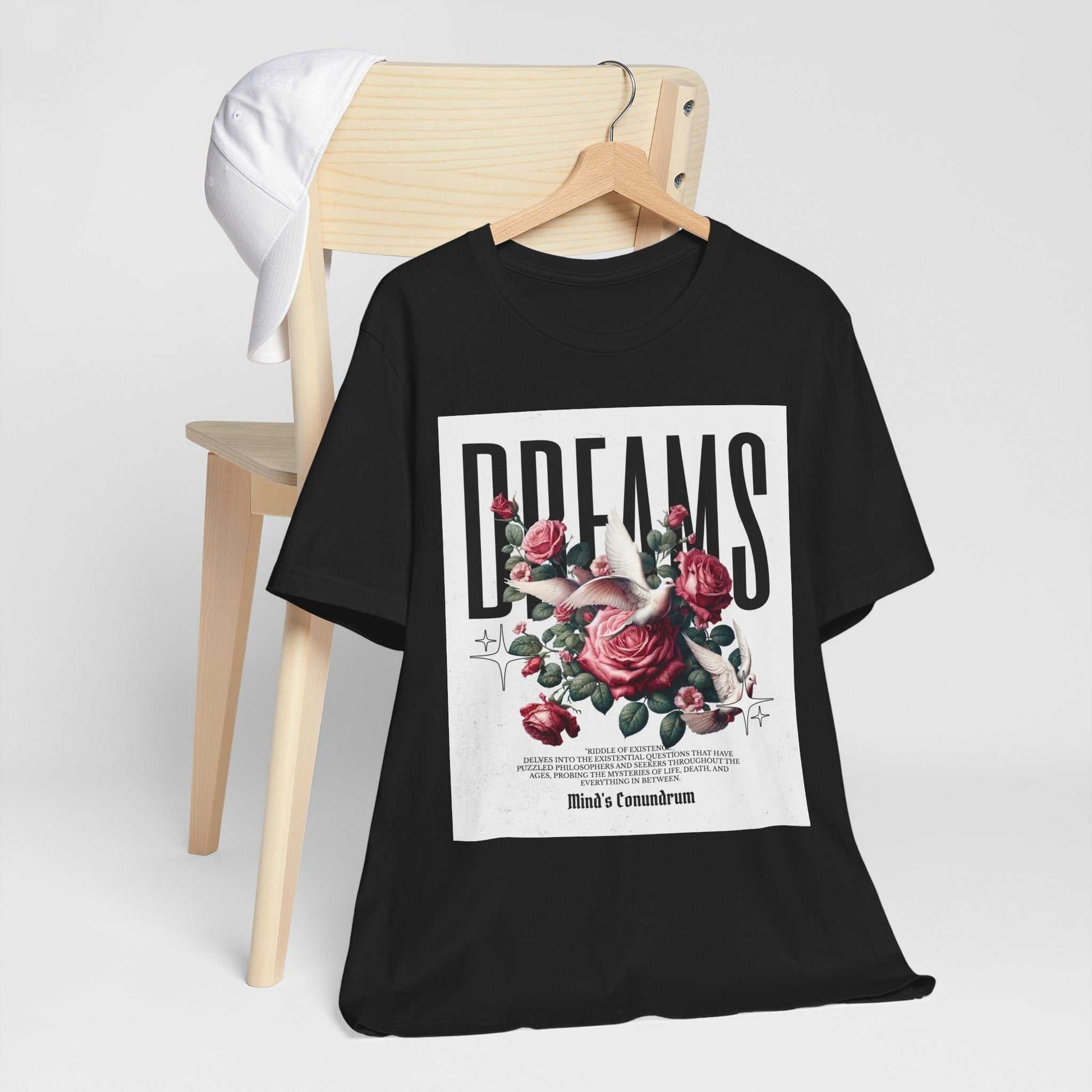 Black Dreams Floral Tee with pink roses and bold typography, hanging on wooden hanger with white cap on chair
