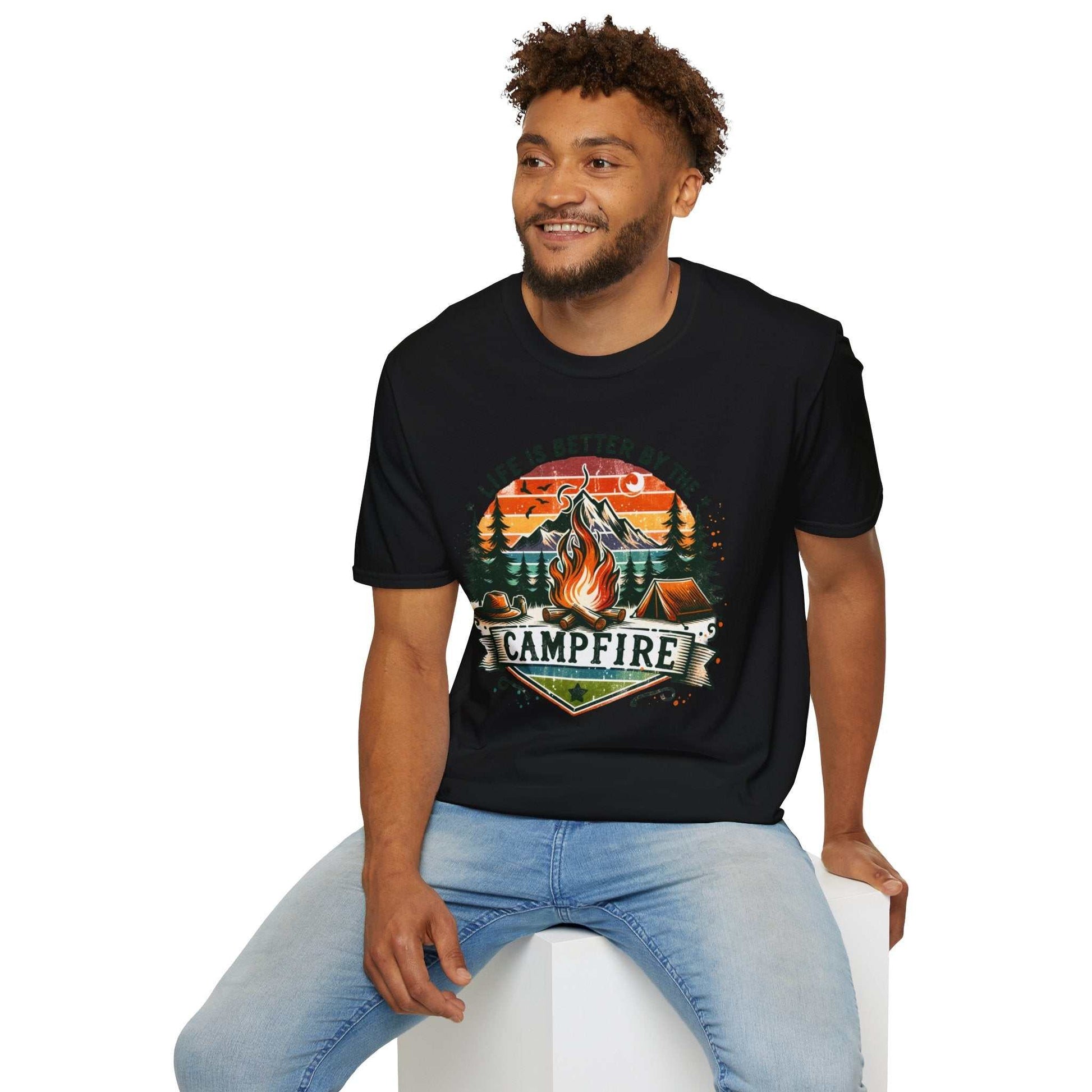 Man wearing black Camping Adventure Unisex T-Shirt with campfire and mountain design sitting on white block