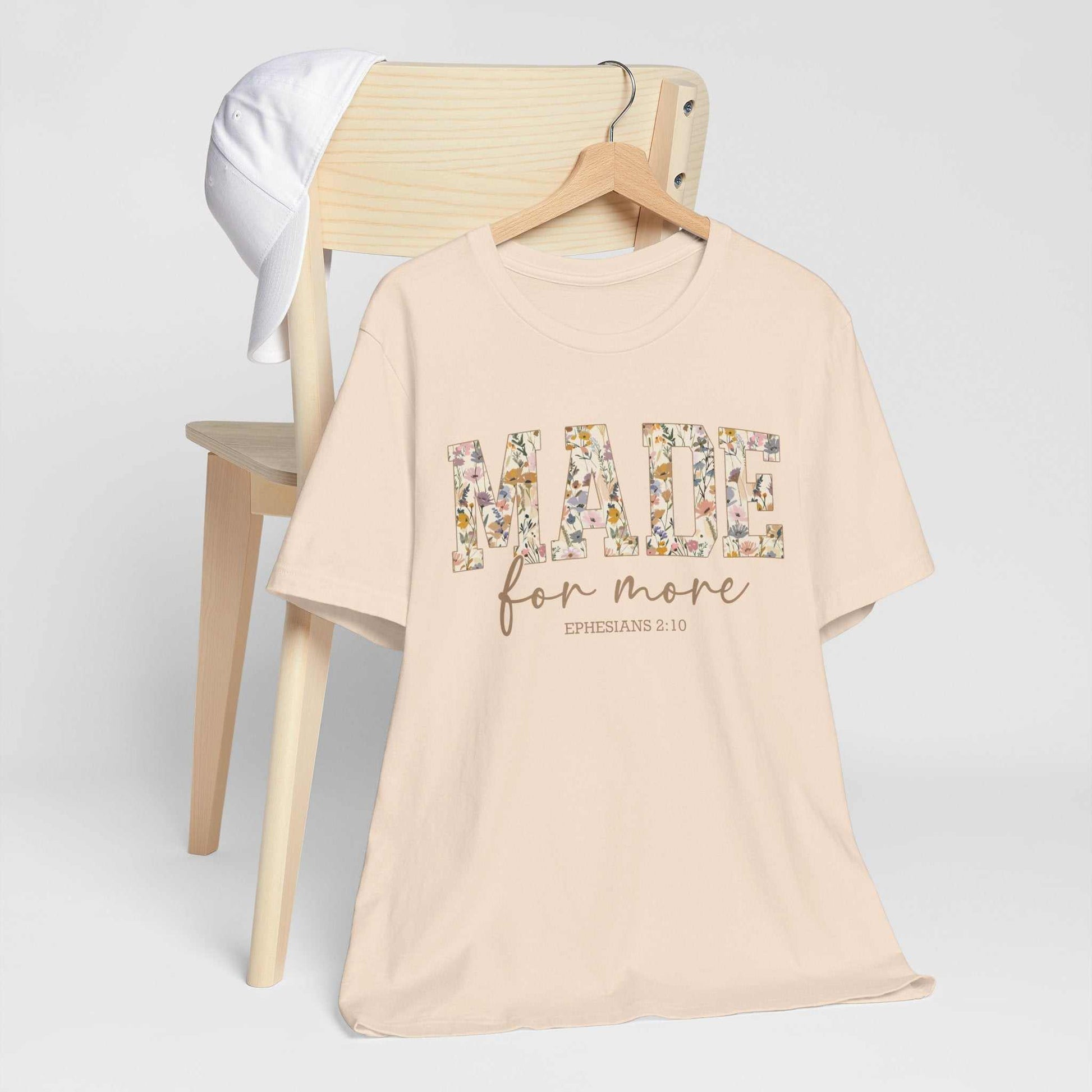 Beige Made for More Tee with floral text design hanging on wooden chair with white cap behind it
