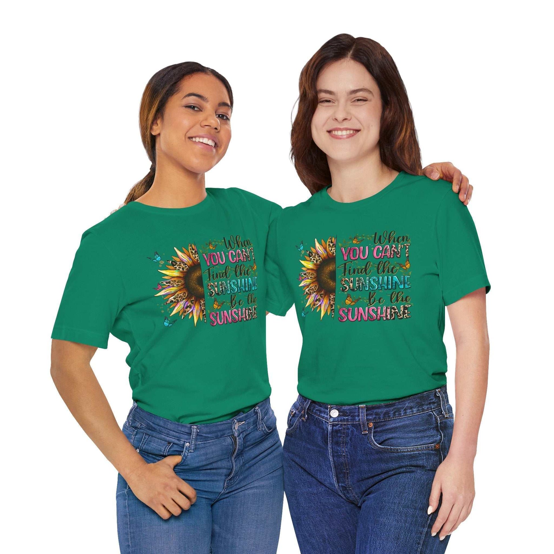 Two women smiling wearing green Sunshine Quote Tee shirts with colorful sunflower and inspirational quote design, styled casually with jeans.