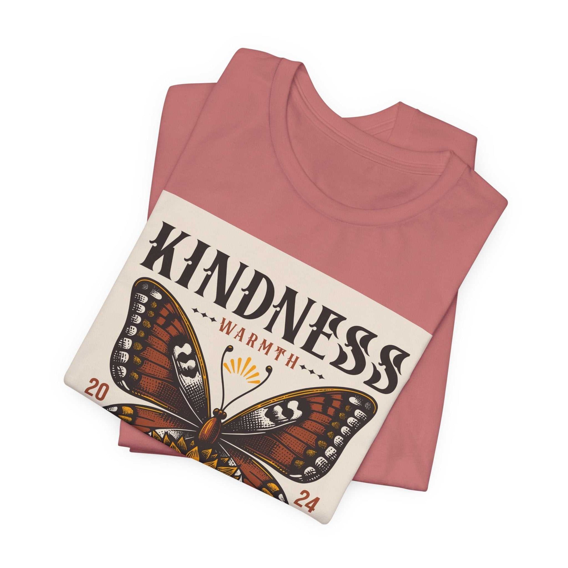 Folded rose pink Kindness Butterfly Tee with butterfly and sunflower design and "Kindness Warmth" text.