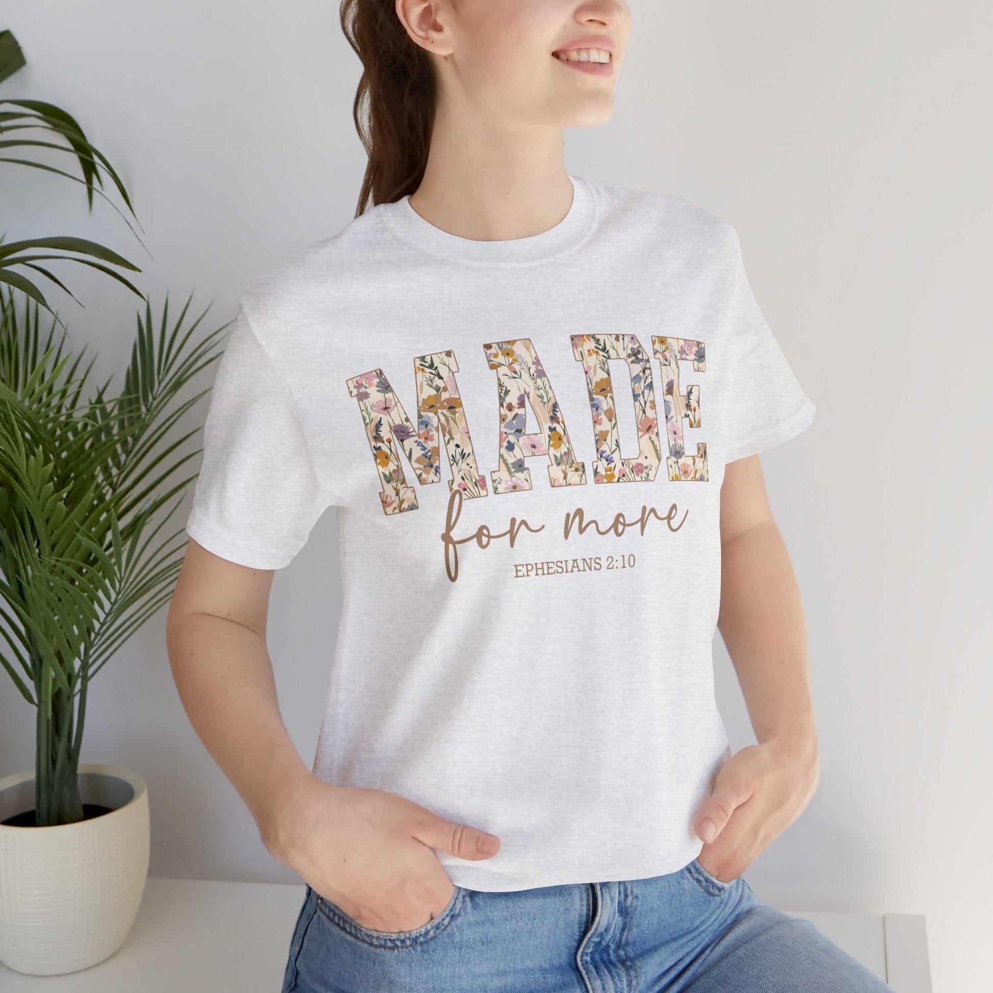 Woman wearing a white Made for More Tee with floral text design and Ephesians 2:10 scripture, paired with blue jeans.