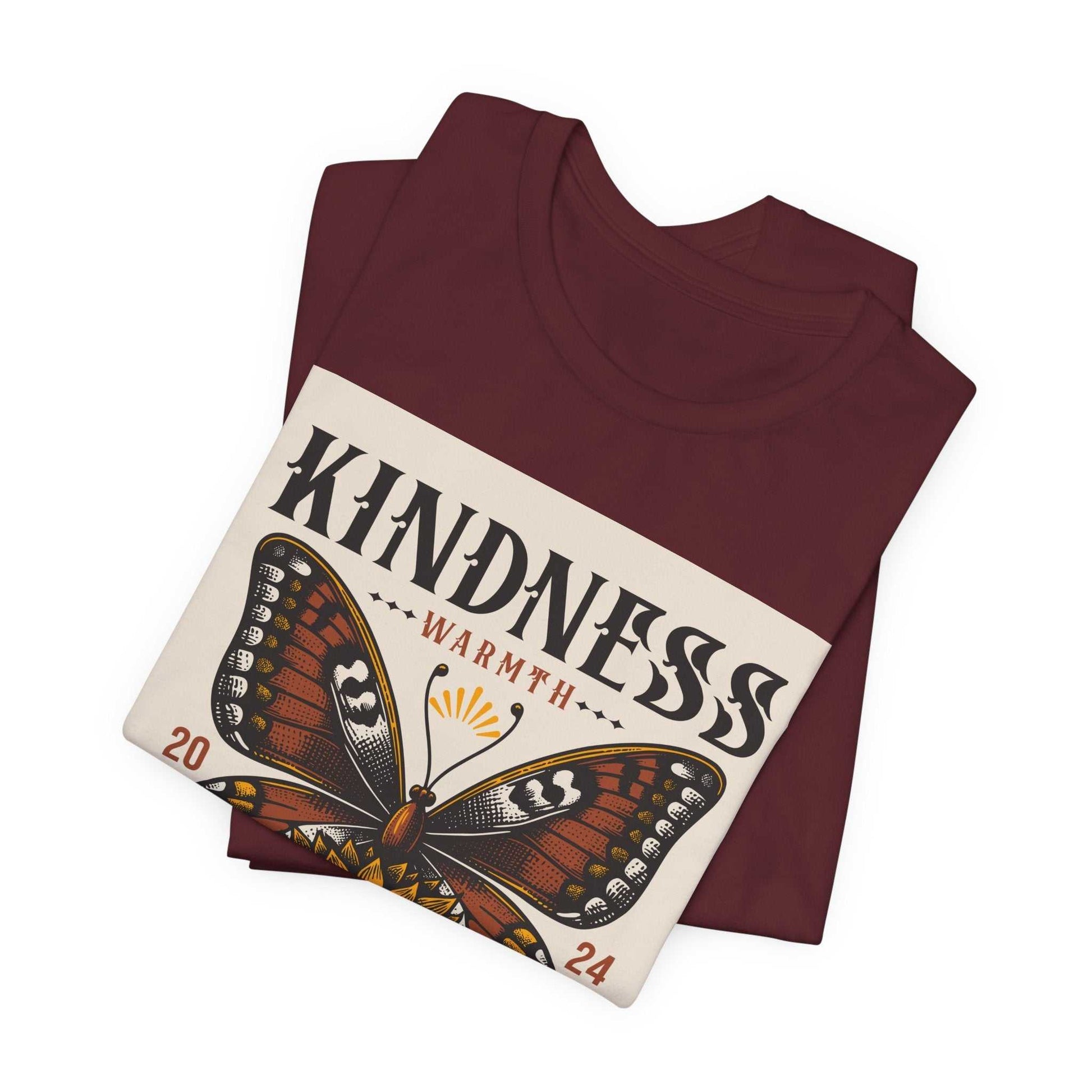 Folded maroon Kindness Butterfly Tee featuring a vibrant butterfly and sunflower design with the word "Kindness" and "Warmth" printed on the front