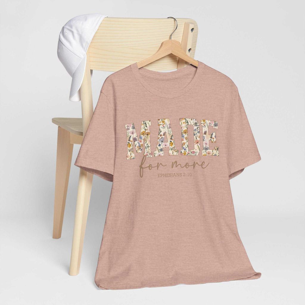 Made for More Tee in dusty pink with floral lettering and inspirational text, displayed on a wooden chair with a white cap hanging on the backrest.