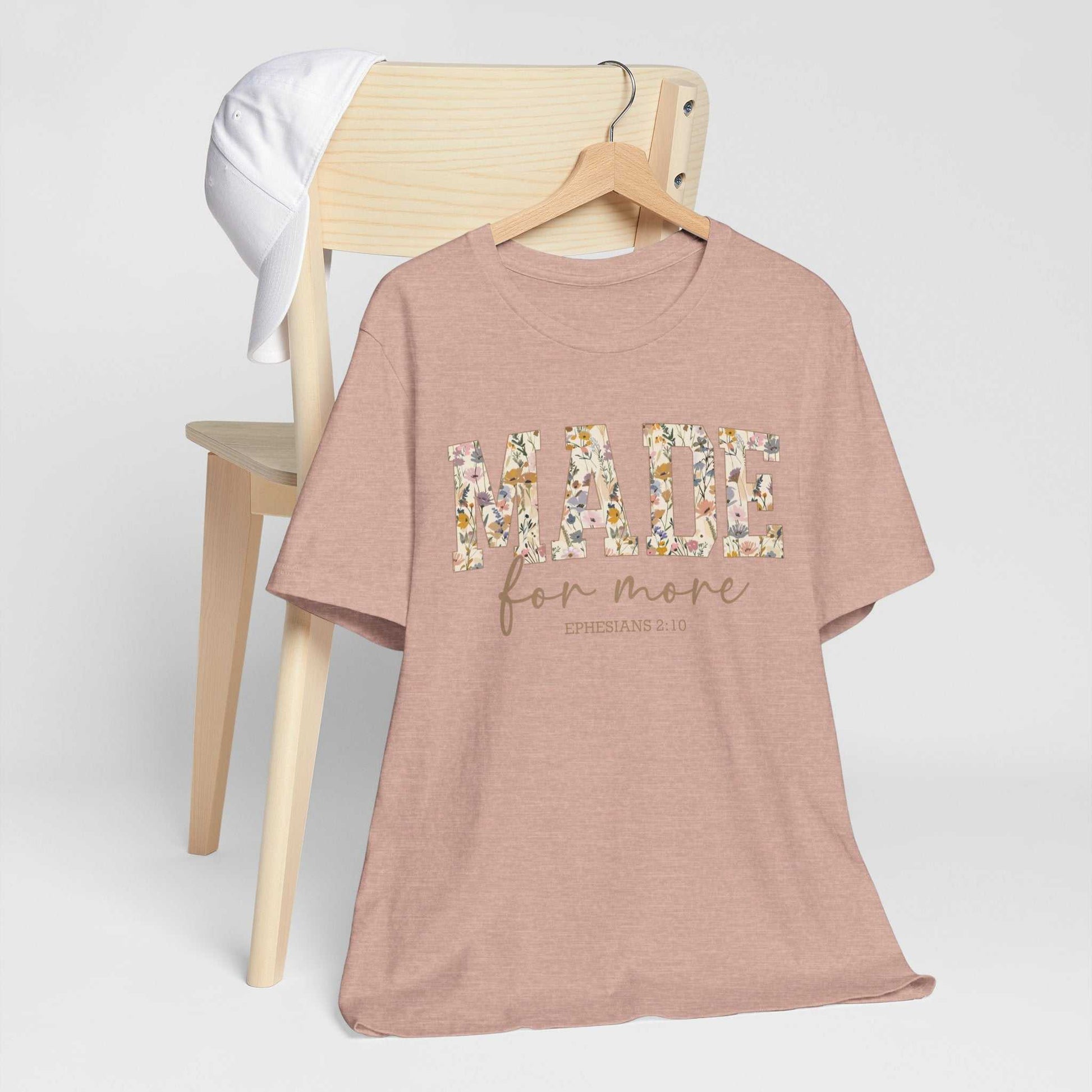 Made for More Tee in dusty pink with floral lettering and inspirational text, displayed on a wooden chair with a white cap hanging on the backrest.