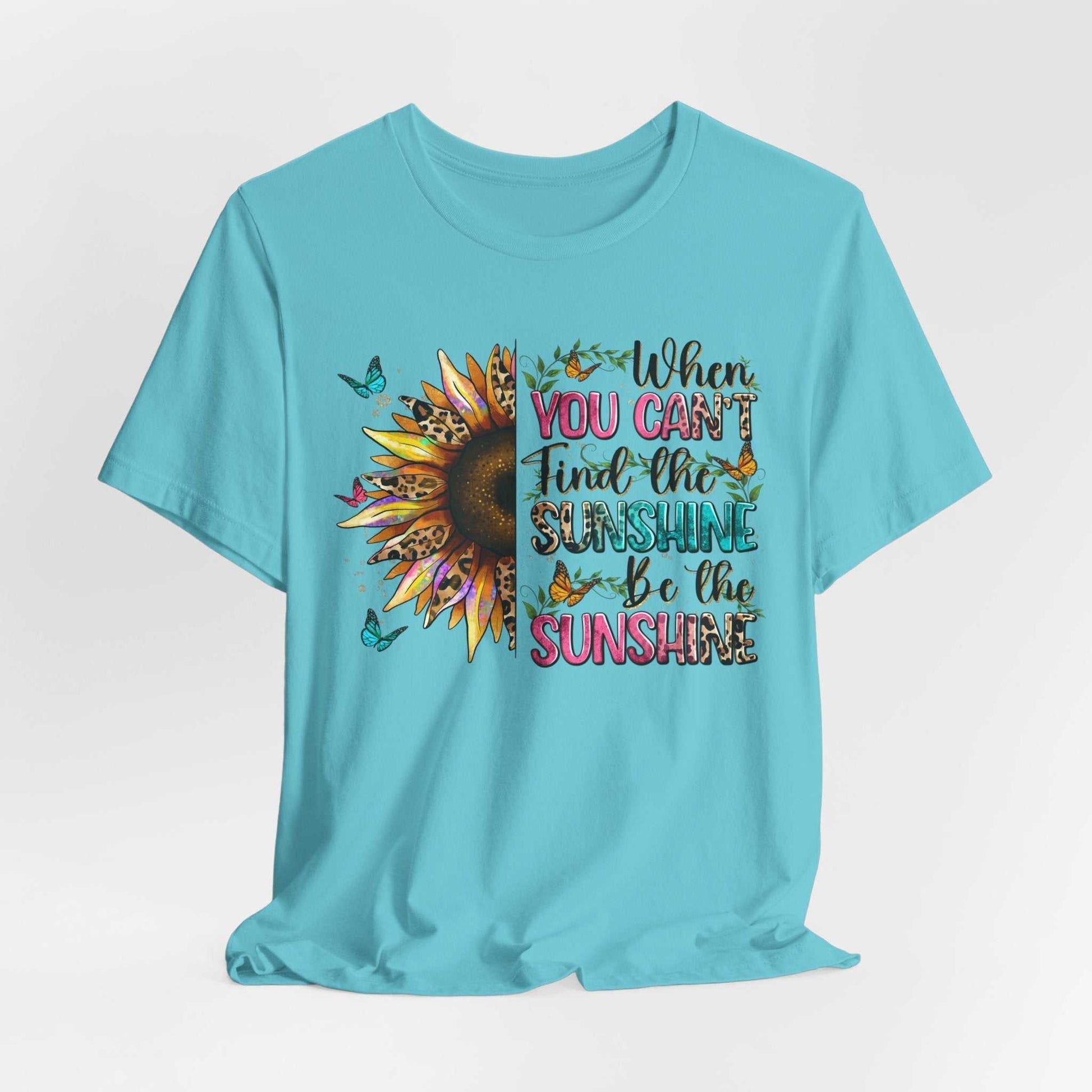 Light blue Sunshine Quote Tee with vibrant sunflower graphic and motivational text "When you can't find the sunshine be the sunshine" surrounded by butterflies